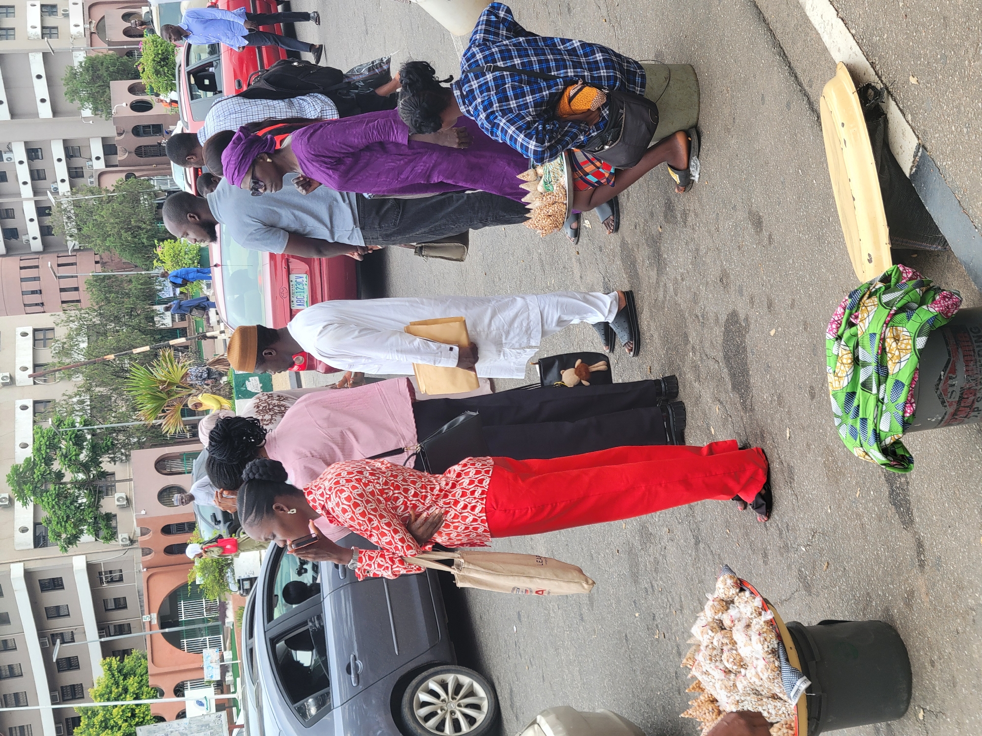 This image depicts a bustling daytime street scene in Abuja, Nigeria, capturing a line of approximately ten to twelve individuals, mostly adults, waiting or queuing along a roadway. The setting appears to be an urban environment, with multi-story buildings and visible air conditioning units lining the street in the background.
In the foreground, a woman wearing a red top and red trousers stands, looking downwards, holding a light brown paper bag that has "LEA" partially visible on its side. Behind her, a diverse group of people in various attires, including casual wear and traditional Nigerian garments like a white agbada and a vibrant purple outfit, form a queue.
To the right, a woman in a blue and white checkered shirt kneels on the ground next to a black bucket filled with various packaged snacks, such as peanuts and popcorn, suggesting she is a street vendor. Another black bucket, adorned with a green, patterned fabric, is also on the ground, along with a yellow, flat object that resembles a broken board.
Several cars are present: a red sedan, clearly displaying a Nigerian license plate "ABC-723BY", is parked in the middle ground, and a dark grey car is closer to the foreground. A traffic barrier or gate is visible further down the street. The lighting suggests an overcast or slightly cloudy day, providing diffused illumination. The scene reflects typical daily life in a Nigerian city, blending formal structures with informal roadside commerce.