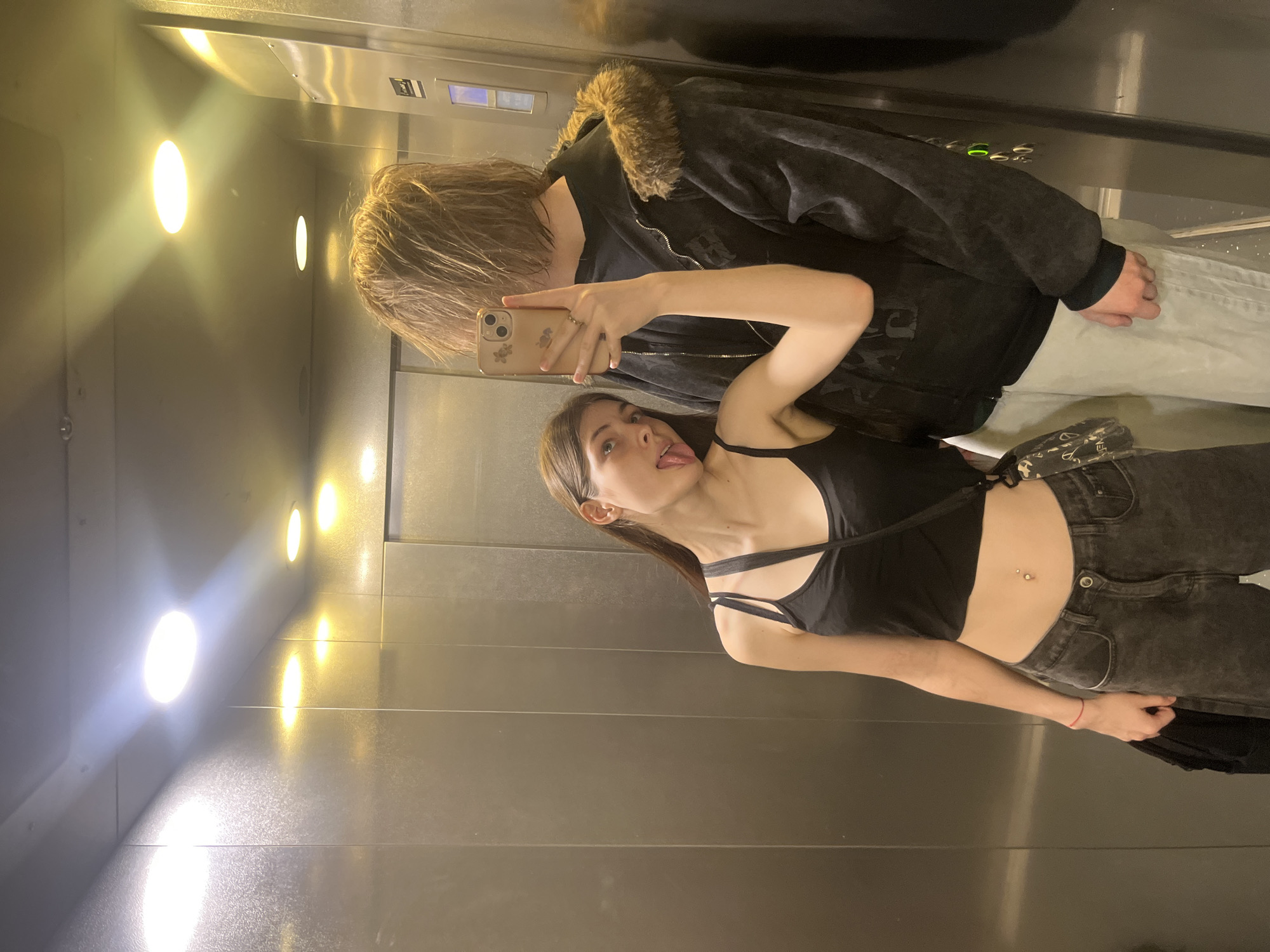 Two young individuals are captured in a selfie taken inside an elevator in Saint Petersburg, Russia.
The female subject, positioned on the left, is a young adult with long, straight, dark brown hair. She is wearing a black spaghetti-strap crop top, revealing her midriff, which features a silver navel piercing. Her lower attire consists of dark-wash jeans. A thin red string bracelet is present on her left wrist, and a silver ring is on her right ring finger. She holds a light-colored smartphone, equipped with a floral-patterned case, in her right hand to take the photograph. Her left hand clutches a dark, folded garment. Her head is tilted slightly to the left, and her tongue is partially extended from her mouth. Her right elbow rests on the left shoulder of the male subject beside her.
The male subject, positioned on the right, is largely obscured by the smartphone. He has shaggy, medium-length, light brown hair that covers his face from the camera's perspective. He is wearing a dark, possibly charcoal-colored, zip-up hooded jacket adorned with a prominent brown fur trim around the hood. Subtle faded patterns are visible on the jacket's fabric. His lower attire consists of light grey or off-white trousers. His right hand is visible at his side.
The elevator interior features reflective, vertically paneled metal walls. The ceiling contains several recessed circular light fixtures, emitting a mix of white and yellowish light that reflects off the metallic surfaces. An elevator control panel with illuminated buttons, one displaying a green light, is partially visible on the right wall behind the male subject.