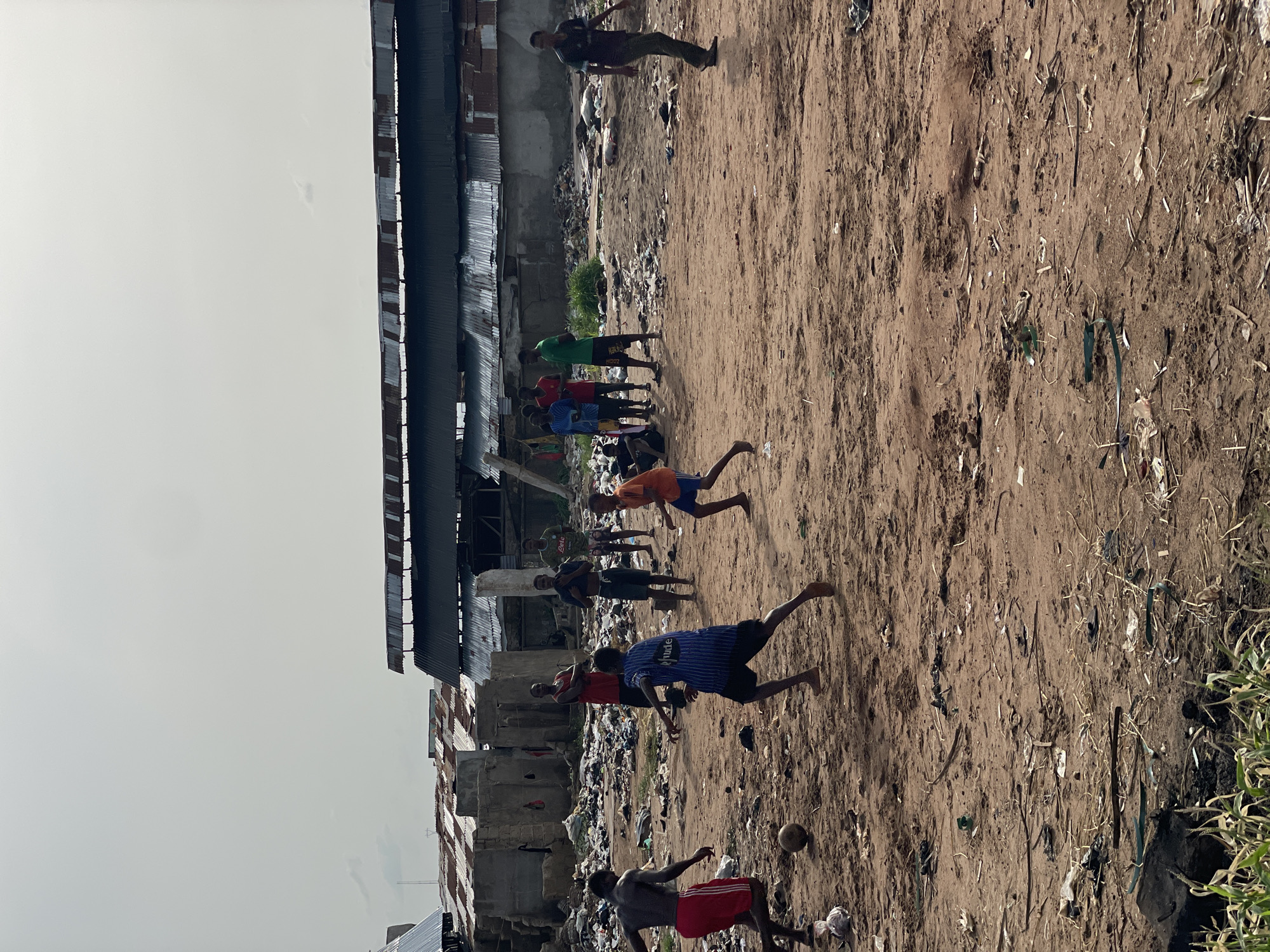 The image depicts a group of people, predominantly young males, playing soccer on a dirt field in what appears to be Onitsha, Nigeria. The playing area is characterized by sparse vegetation and scattered debris, suggesting a marginal or undeveloped space. Adjacent to the field is a densely built-up area consisting of corrugated metal structures and buildings with visible signs of deterioration. Litter is present throughout the scene. The sky is overcast, providing soft, ambient light.