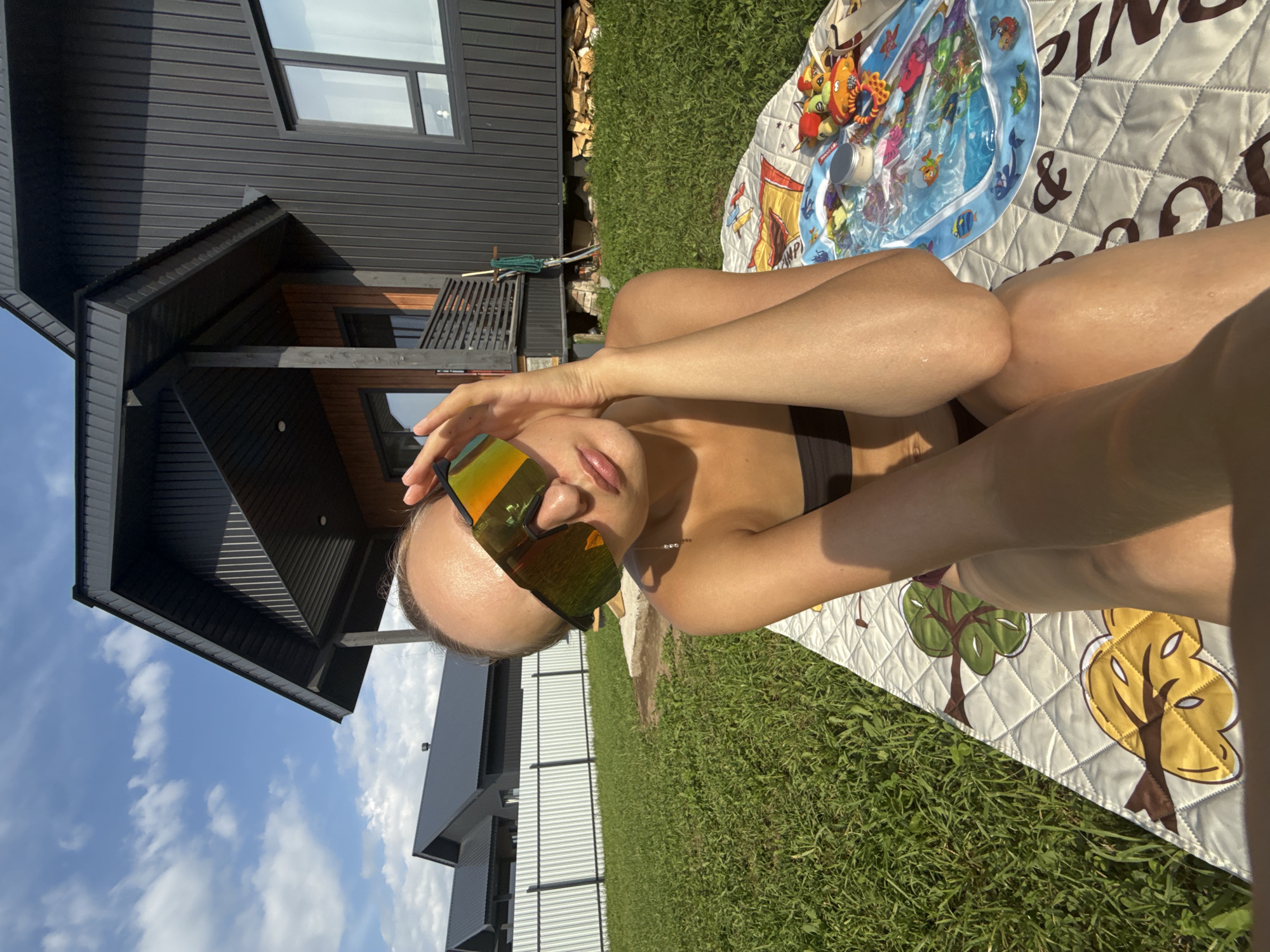 A person is lying on their back on a quilted blanket spread out on green grass. The person is wearing sunglasses with mirrored lenses that reflect the surroundings. The person's arms are bent and resting on their chest and stomach, and their legs are bent at the knees. Next to the person on the blanket is a baby water play mat with toys and a small jar. In the background, there is a modern black house with large windows, and a blue sky with white clouds. The sun is shining, indicating it is daytime. The scene appears to be a backyard or outdoor recreational area. The overall mood is relaxed and summery. There is no visible text in the image.