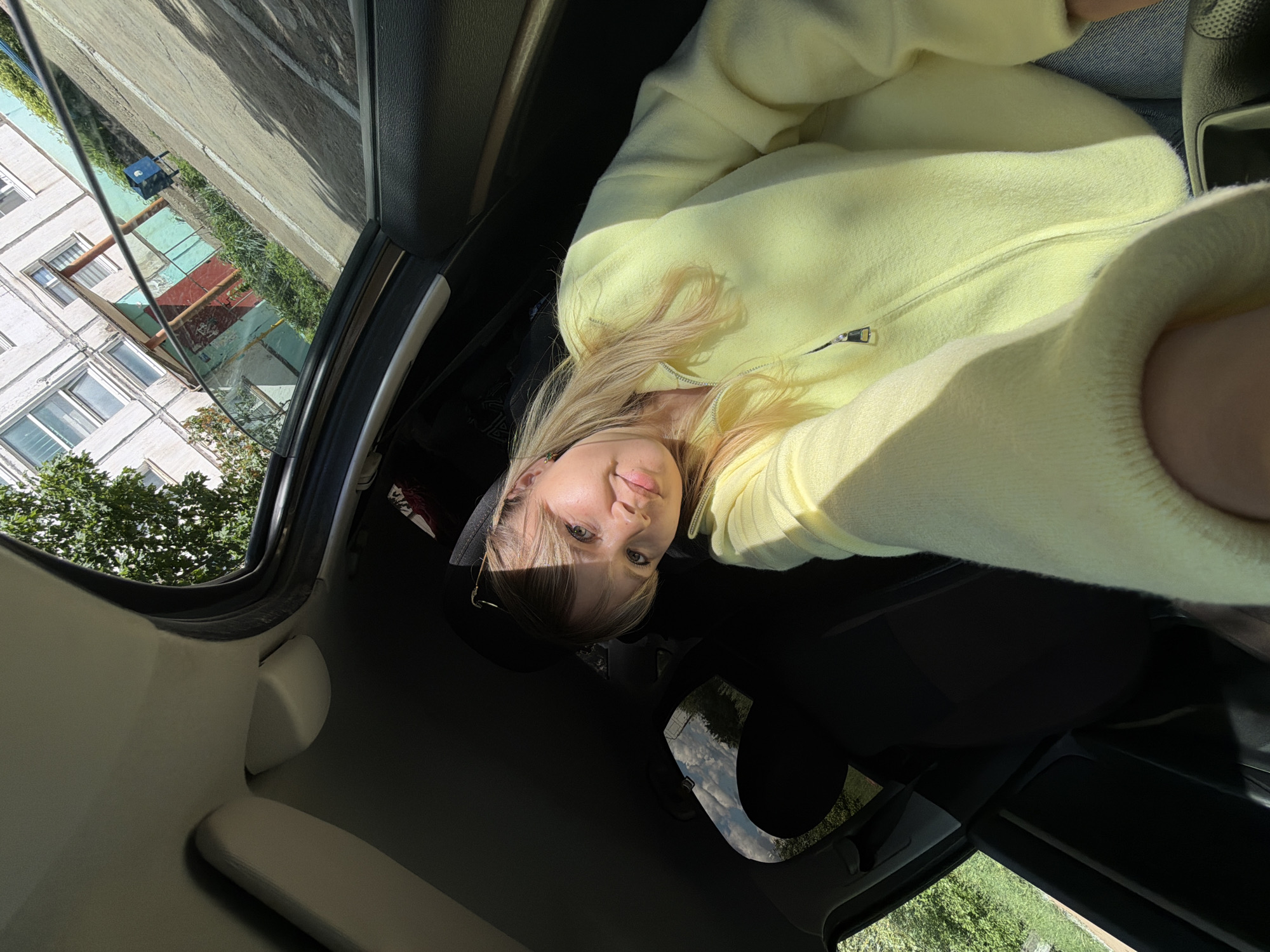 A woman with blonde hair is photographed from inside a vehicle, possibly a car. She is wearing a pale yellow sweater. The vehicle's sunroof is open, providing a view of an apartment building with multiple windows, trees, and a sidewalk outside. A glimpse of the outside environment shows green foliage, suggesting an urban area in Chaikovsky, Russia, based on the request. The overall impression is one of casual travel within a residential area.
