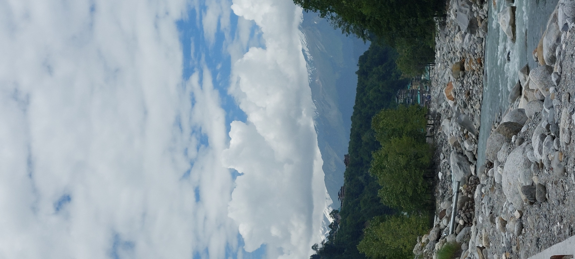 The image depicts a scenic view of the Beas River flowing through Kalath, India. In the foreground, the riverbed is filled with a multitude of large and small grey and white rocks. The water is a murky blue-green and appears to be flowing rapidly over the rocks. Dense green trees line the banks of the river, and in the background, a mountain range with snow-capped peaks is visible. The sky is a pale blue, dotted with fluffy white clouds. Several buildings, possibly hotels or guesthouses, are nestled on the slopes of the mountains. The overall impression is one of natural beauty and tranquility.