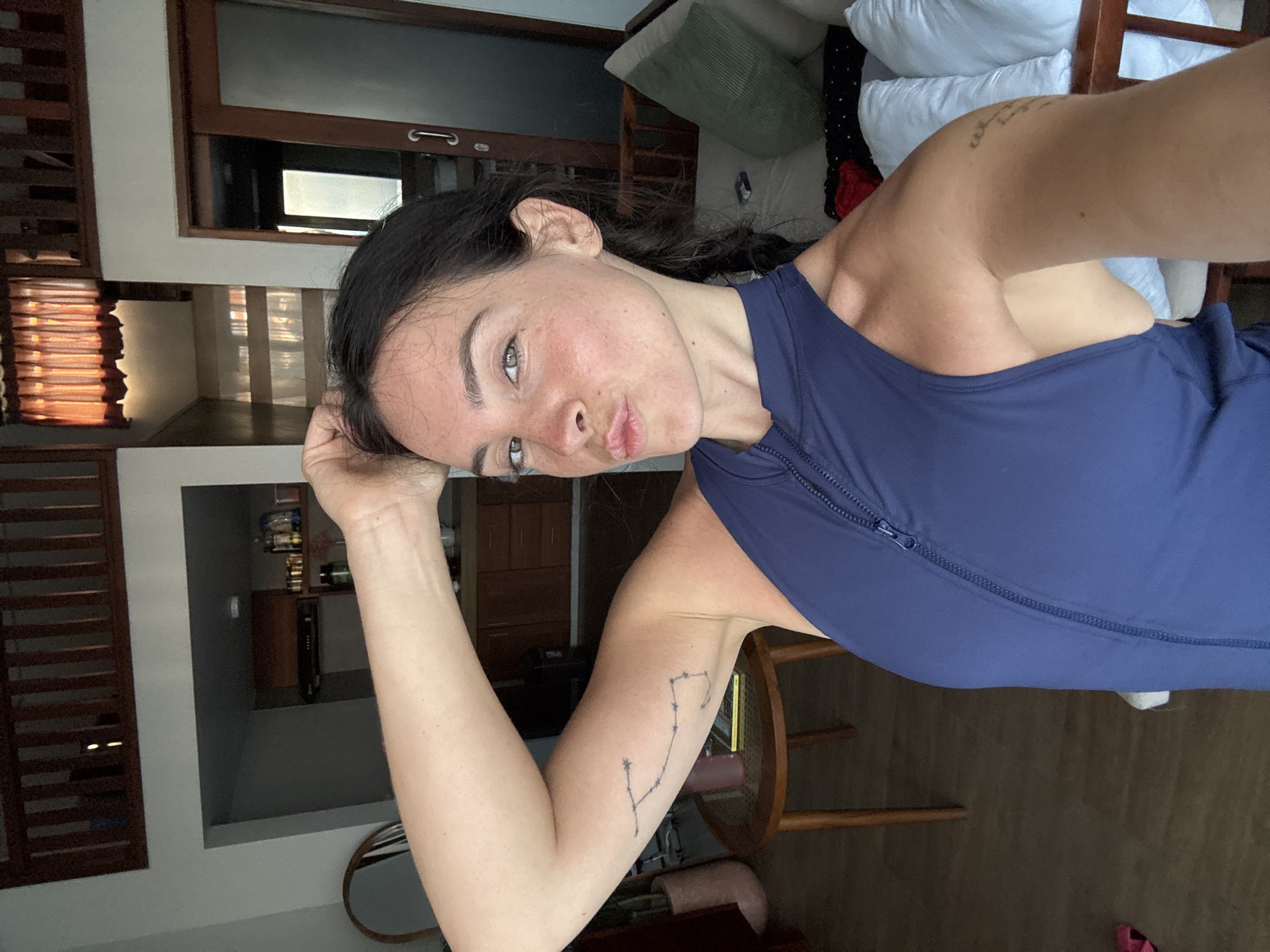 A woman is taking a selfie in what appears to be a hotel room in North Kuta, Indonesia. She is wearing a dark blue sleeveless top with a zipper. The room has wooden floors and furniture, with a kitchen area visible in the background. There is also a seating area with pillows and a window with orange curtains. The woman has dark hair pulled back in a braid and is making a puckered face towards the camera. She has tattoos on her arm.