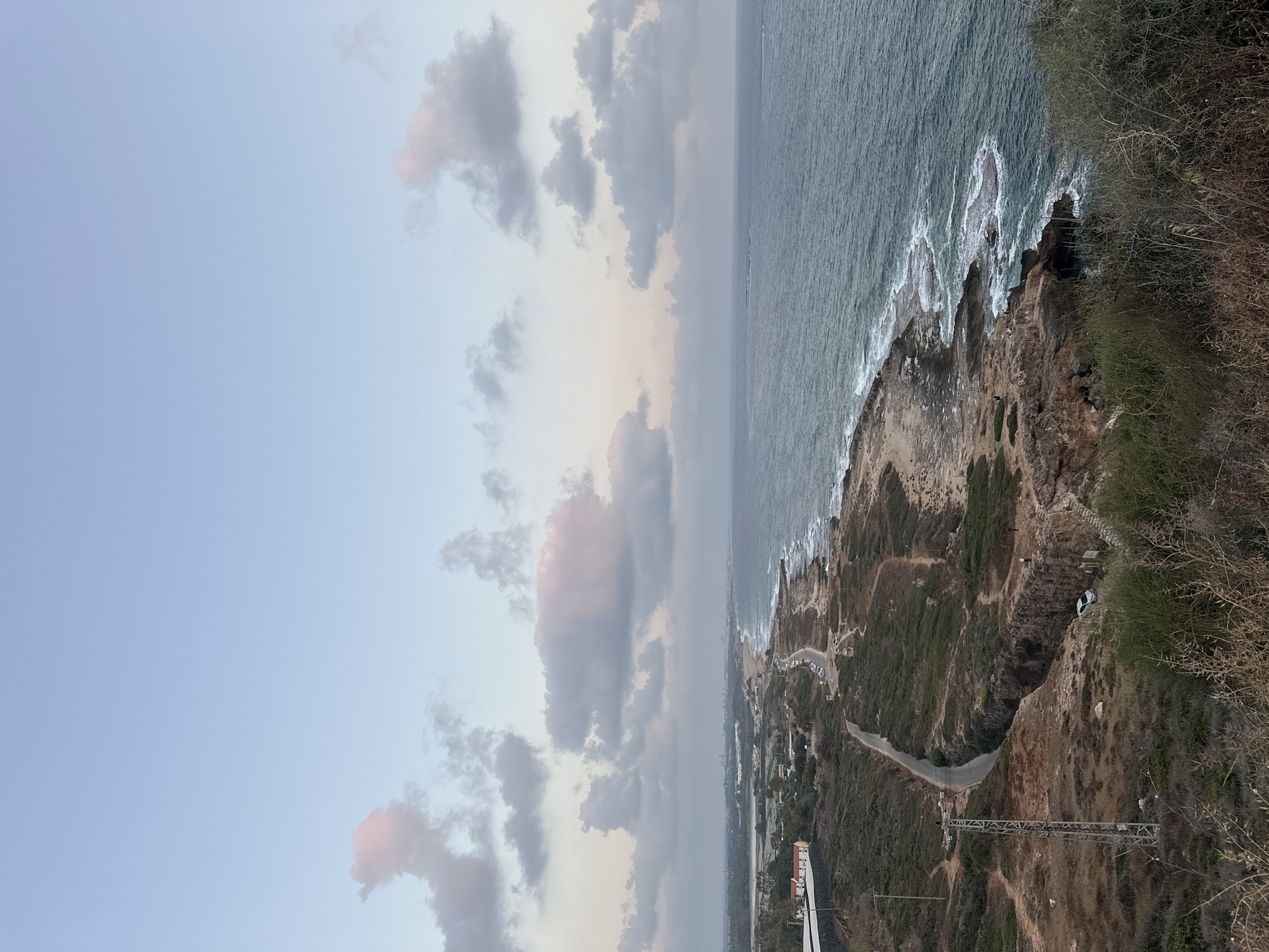 An elevated, wide-angle, outdoor, daylight shot captures a coastal landscape in the city of Mate Asher Regional Council, Israel. The sky, a soft blue with scattered clouds tinged with pink, dominates the upper portion of the image. Below, the deep blue sea meets a rugged coastline of rocky outcrops and sandy beaches. A winding road snakes along the terrain, with sparse vegetation dotting the hillsides. In the distance, a city skyline is visible, hinting at urban development. A few cars are parked along the road, and a handful of people are seen near the coastline. The scene suggests a serene, natural environment juxtaposed with human presence.