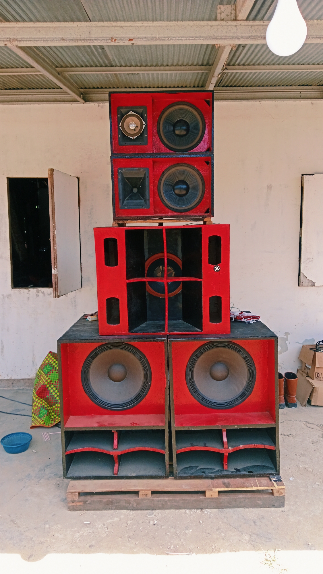 The image depicts a stack of large, red speaker cabinets outdoors. The speakers are arranged in three tiers. The top tier consists of two smaller speaker units, each with a tweeter and a woofer. The middle tier is a larger, open-fronted cabinet with visible internal divisions and a central circular element. The bottom tier comprises two substantial speaker cabinets, each with a large woofer. The speaker units are placed on a wooden pallet on a concrete surface. To the left of the speakers, there is a colorful patterned bag and a blue bowl. In the background, a white building with open windows is visible, and a corrugated metal roof extends over the area. A light bulb hangs from the ceiling. To the right, cardboard boxes and a pair of brown boots are present. The scene appears to be in an outdoor, possibly semi-constructed or storage area, under a roofed structure. The location is identified as Karang Indah, Indonesia.