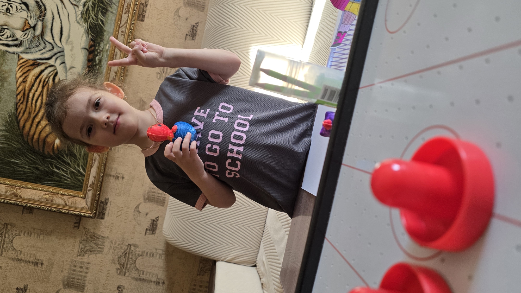 A young girl with light brown hair pulled back is standing in front of a couch in what appears to be a living room. She is wearing a dark gray t-shirt with pink trim on the collar and sleeves. The t-shirt has pink text that reads "HAVE TO GO TO SCHOOL." In her right hand, she holds a red and blue toy, and her left hand is raised, displaying the peace sign. Behind her, a framed painting of a tiger hangs on the wall, which is decorated with a patterned wallpaper. In the foreground, a portion of an air hockey table is visible, with two red paddles in focus. The scene is brightly lit, suggesting daylight. The location is identified as Верхнеказымский, Russia.