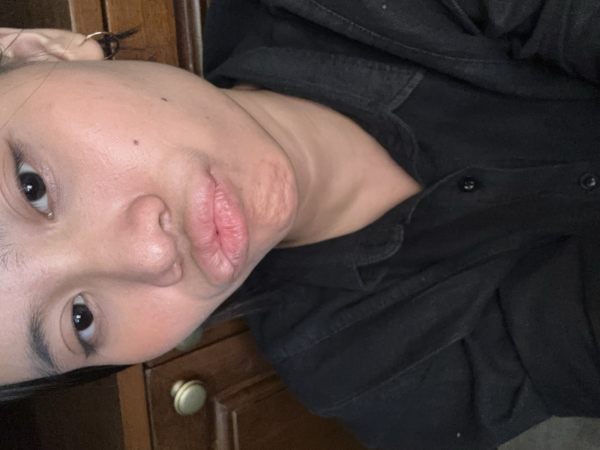 The image is a close-up, selfie-style photograph of a young person's face, from the nose down to the collarbone. The subject is wearing a black collared shirt. The background appears to be indoors, with a dark wooden piece of furniture, possibly a dresser or cabinet, visible to the left and right. A light-colored drawer knob is visible on the left. The lighting is dim. The overall impression is that this photograph was taken in a residential setting. No specific location in South Jakarta, Indonesia, can be identified from the visual information provided.