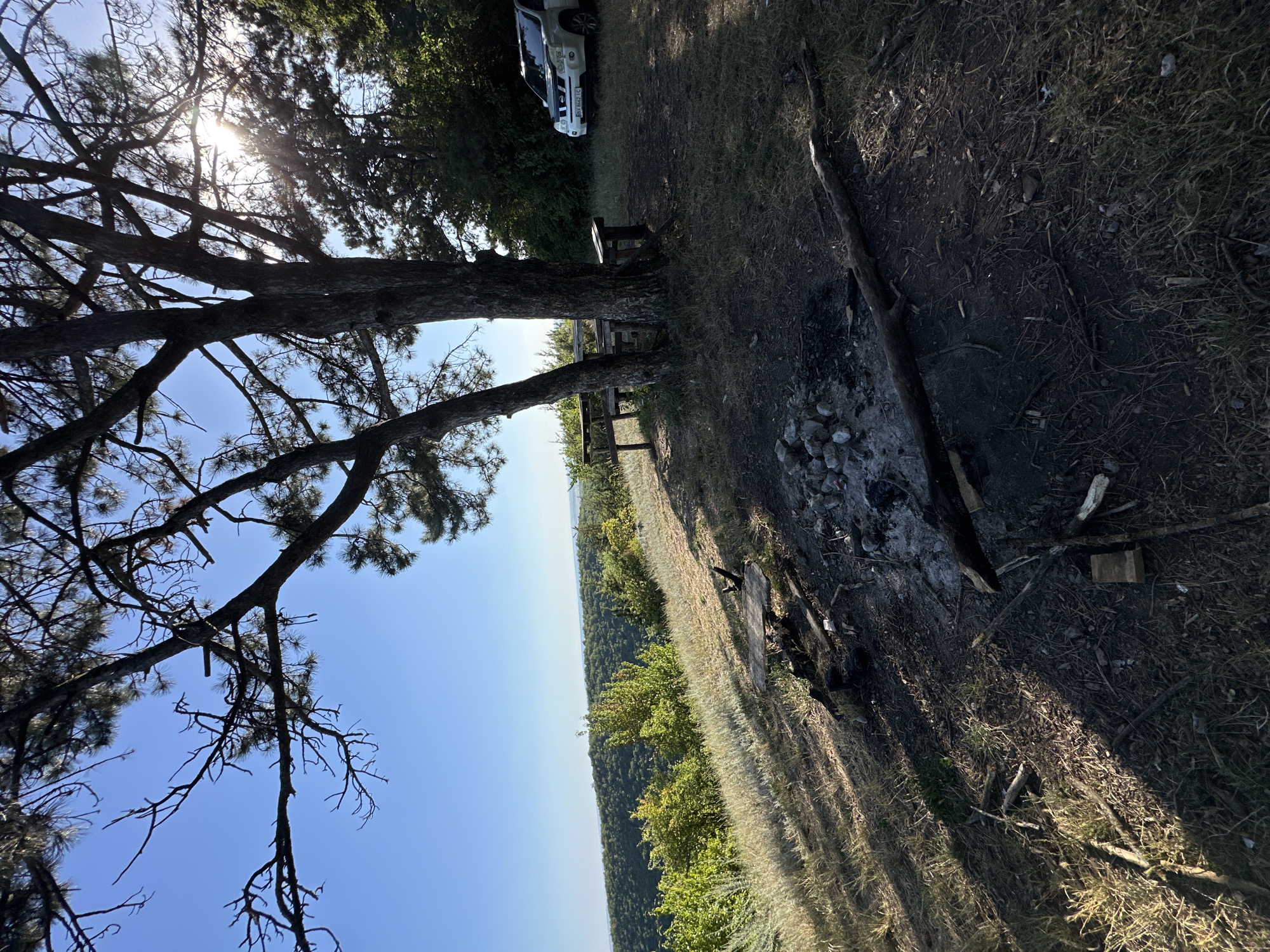 The image depicts an outdoor campsite located on a hill with a view of a forested valley and a distant horizon under a clear blue sky. A large tree with bare branches casts a shadow over a picnic table and benches. In the foreground, a fire pit with scattered rocks and debris is visible. A white SUV is parked to the right of the picnic area. The scene appears to be in Pryvorottia Druhe, Ukraine, with the natural landscape dominating the view. The overall impression is one of a rustic, natural setting, possibly a recreational spot.