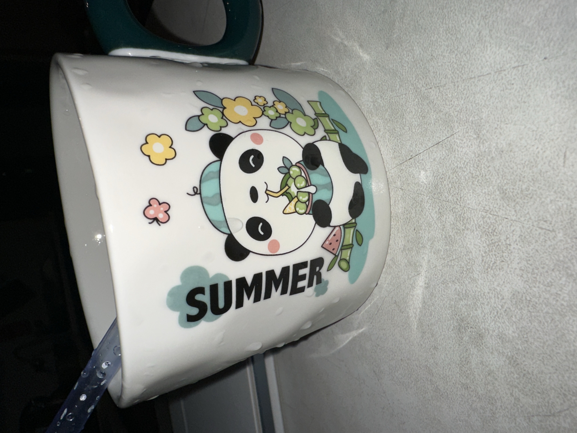 A white mug with a cartoon panda and the word "SUMMER" is on a speckled white surface. The mug has water droplets on its exterior. A blue spoon is in the mug.
