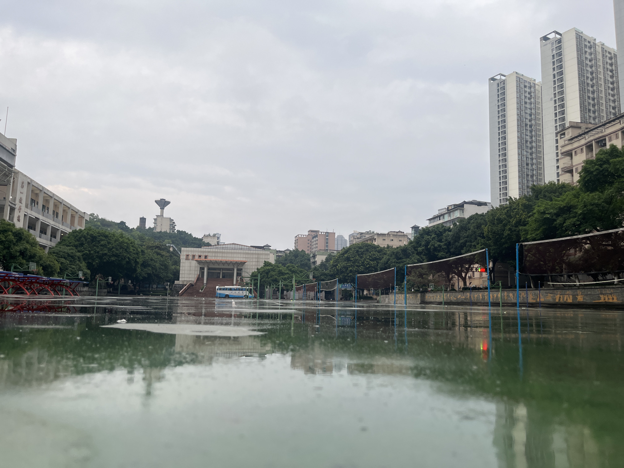 This image depicts a waterlogged outdoor athletic court in Yuzhong District, China, under an overcast sky. The foreground is dominated by a reflective, green-hued surface of standing water, indicating recent rainfall, with visible ripples from raindrops.
In the midground, from left to right:
* Red metal structures, possibly bleachers or sports equipment frames, are partially submerged in the water on the far left.
* A light blue and white bus is parked near the base of a building with prominent exterior stairs. This building has a brown tile roof and beige walls, suggesting a public building like a gymnasium or assembly hall.
* Further to the right, a series of dark brown nets supported by blue poles are erected across what appears to be a volleyball court. Behind these nets, a low wall features yellow and blue painted characters or graffiti.
* A traffic light with illuminated red and green signals is visible behind the nets on the right side.
* Lush green trees line the midground, forming a buffer between the court area and more distant structures.
The background reveals a hilly urban landscape characteristic of Yuzhong District:
* On the far left, a multi-story building with a flat roof and numerous windows has Chinese characters "第一中学" (Diyi Zhongxue - First Middle School) inscribed on its facade, identifying it as a school building.
* Centered on a distant hill, a distinctive, tall, inverted-cone-shaped water tower is visible, accompanied by another darker, slender tower structure.
* The right side of the background is dominated by multiple tall, modern high-rise buildings, likely residential, with numerous balconies and windows. These light-colored skyscrapers rise above the tree line and other smaller, older buildings scattered across the hilly terrain.
* The sky is uniformly overcast and light grey, contributing to the diffused lighting of the scene.
No individuals are visible in the image. The scene suggests an unoccupied public or school area after rain.