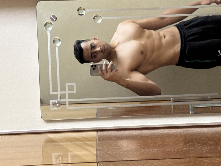 The image displays a shirtless male individual taking a mirror selfie within an indoor setting, consistent with a private residence in Kolkata, India. The male, with a muscular physique, short dark hair, and a slight beard, is positioned to display his upper body. He wears black athletic shorts with a green vertical stripe and a white Puma logo on the left thigh. In his left hand, he holds a silver-colored smartphone, used to capture the image, and a gold-colored bracelet is visible on the same wrist. The subject's expression is neutral, and his gaze is directed towards the phone/camera. The rectangular mirror features decorative etched geometric patterns and four circular reflective embellishments. To the left of the mirror, a section of light brown wooden furniture, characterized by vertical panels and a subtle grain pattern, is visible. The wall reflecting behind the subject in the mirror appears to be a plain, light color.