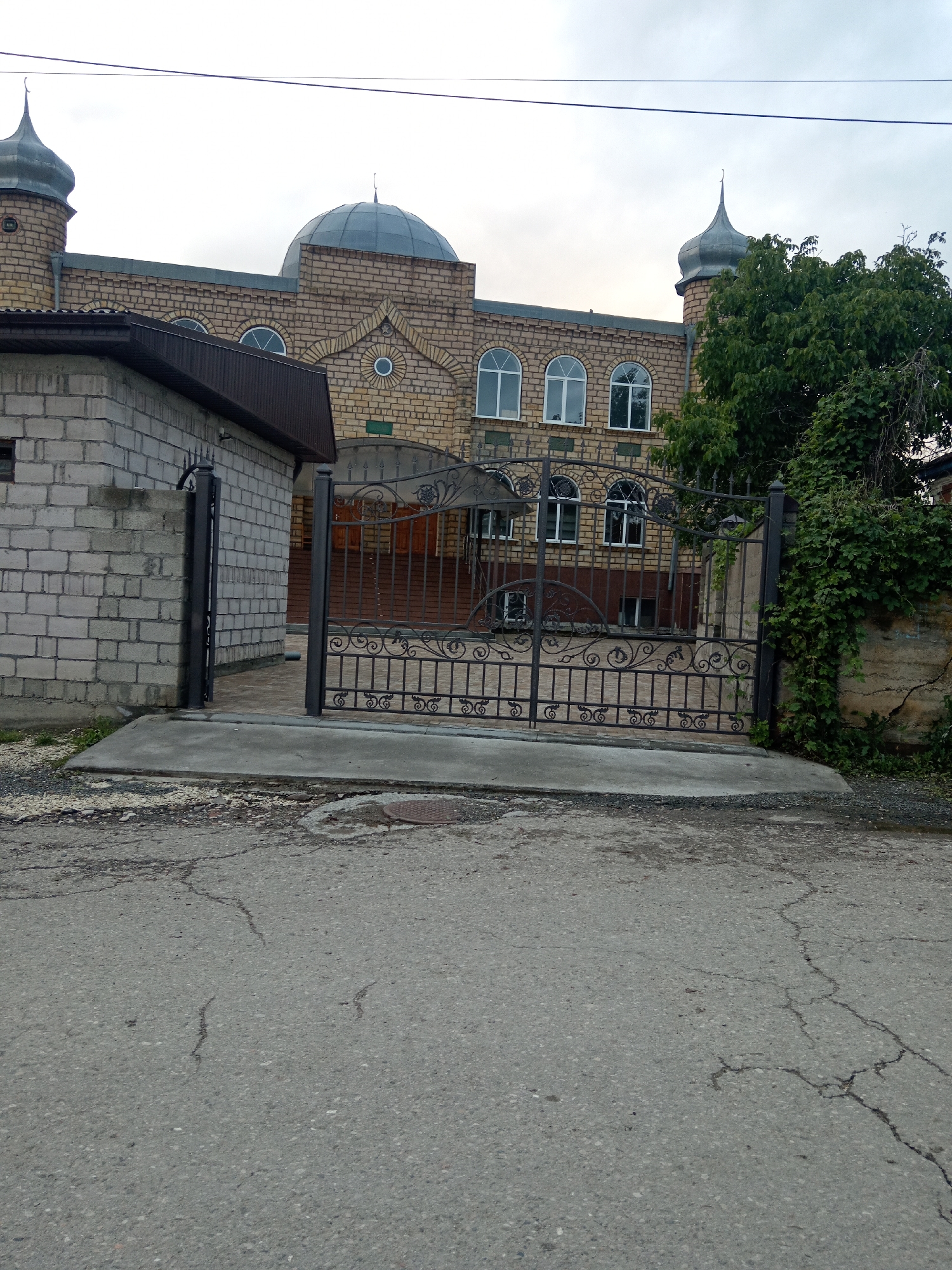 The image depicts a view of a building with arched windows and domes, characteristic of a mosque, located in the city of Uchkeken, Russia. The building is constructed of yellow brick. In the foreground, there is a decorative black metal gate that is partially open, leading into the property. To the left of the gate is a grey brick structure, possibly a wall or a smaller building. The ground in front of the gate and the building is paved, with cracked asphalt on the visible road. Lush green trees and vines are present on the right side of the image, partially obscuring a stone wall. The sky is overcast. There are no people visible in the image.