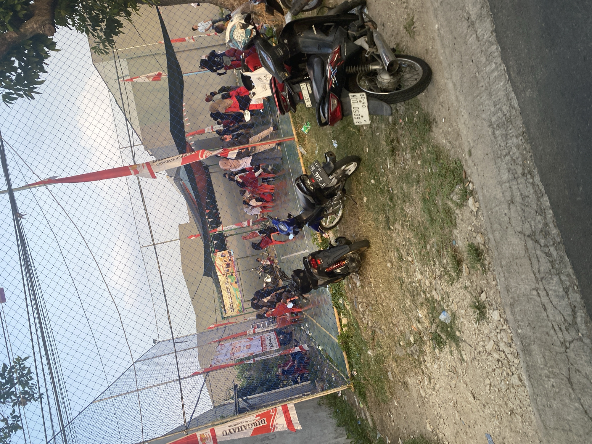 The image depicts an outdoor scene likely in Depok, Indonesia, with a sports court enclosed by a net fence. Several people, including children and adults, are gathered on and around the court, with some seated on red chairs. Red and white Indonesian flags are prominently displayed, suggesting a national event or celebration.
Several motorcycles are parked in the foreground and around the perimeter of the court. The ground is a mix of dirt, gravel, and patchy grass. A concrete barrier runs along the lower edge of the image, adjacent to a paved road. The sky is visible above the net, with some trees and electrical wires also present. The overall impression is a community gathering or event taking place in an urban or semi-urban environment.