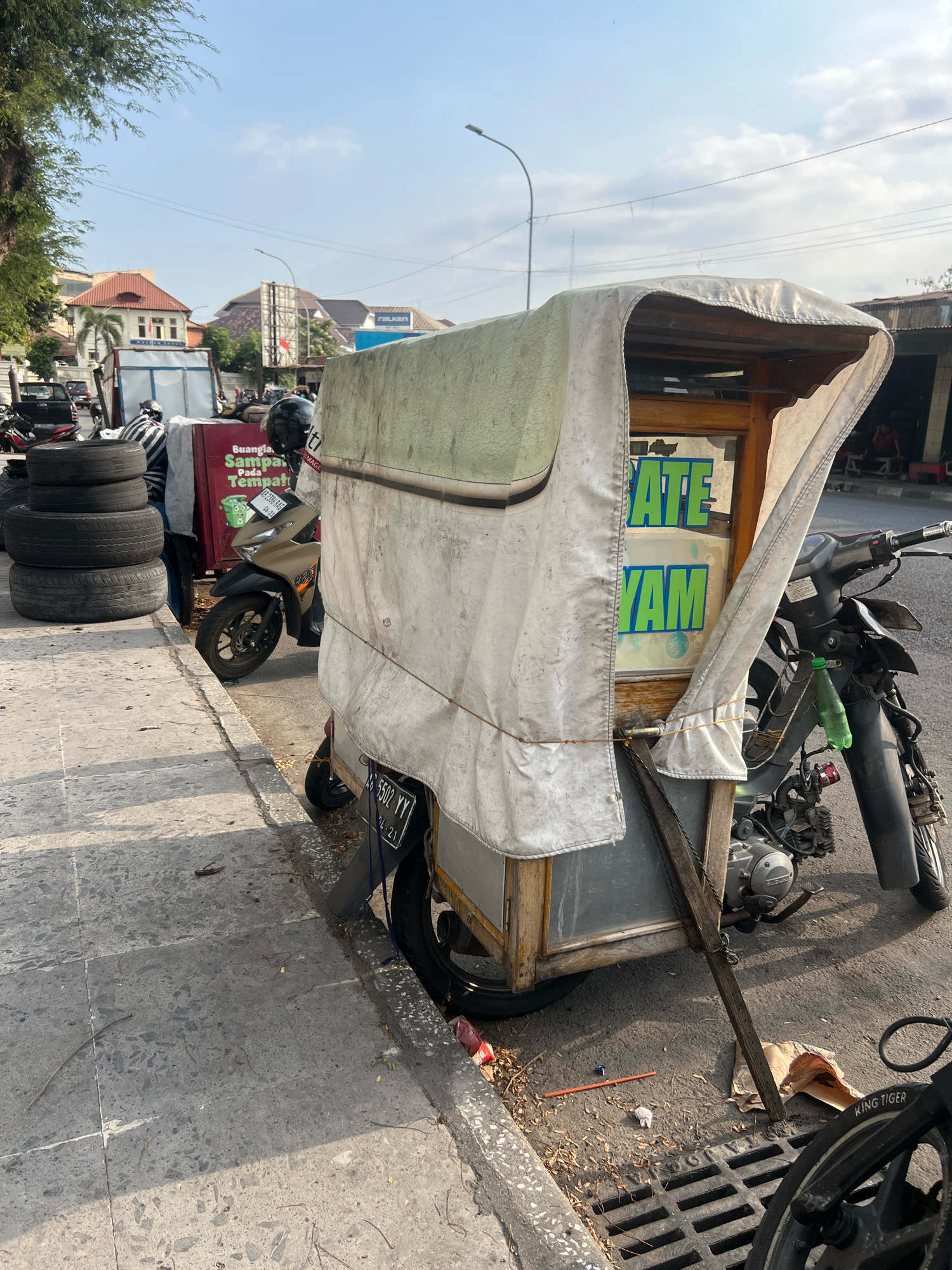 An outdoor, daytime, medium shot captures a food cart, likely a motor-powered vendor, parked on the side of a street in Yogyakarta, Indonesia. The cart, constructed of wood and metal, is covered with a weathered, off-white tarp that drapes over its frame. Visible through a plastic window on the cart are the words "ATE" and "YAM" in bright green lettering, suggesting the type of food sold. The cart is attached to a motorcycle, its rear wheel and engine partially visible. A second motorcycle, a lighter gold color, is parked behind the food cart. To the left of the cart, a stack of four used tires is piled on the sidewalk, and further down the sidewalk, a red cooler with white lettering is visible. The street surface appears to be asphalt, and the sidewalk is made of concrete tiles with a textured pattern. A metal grate, likely a drainage cover, is located on the street in front of the cart. In the background, buildings with red-tiled roofs and other structures are visible, along with trees and a street light. The sky is clear with some wispy clouds. The overall scene depicts a typical street vendor setup in a bustling urban environment.