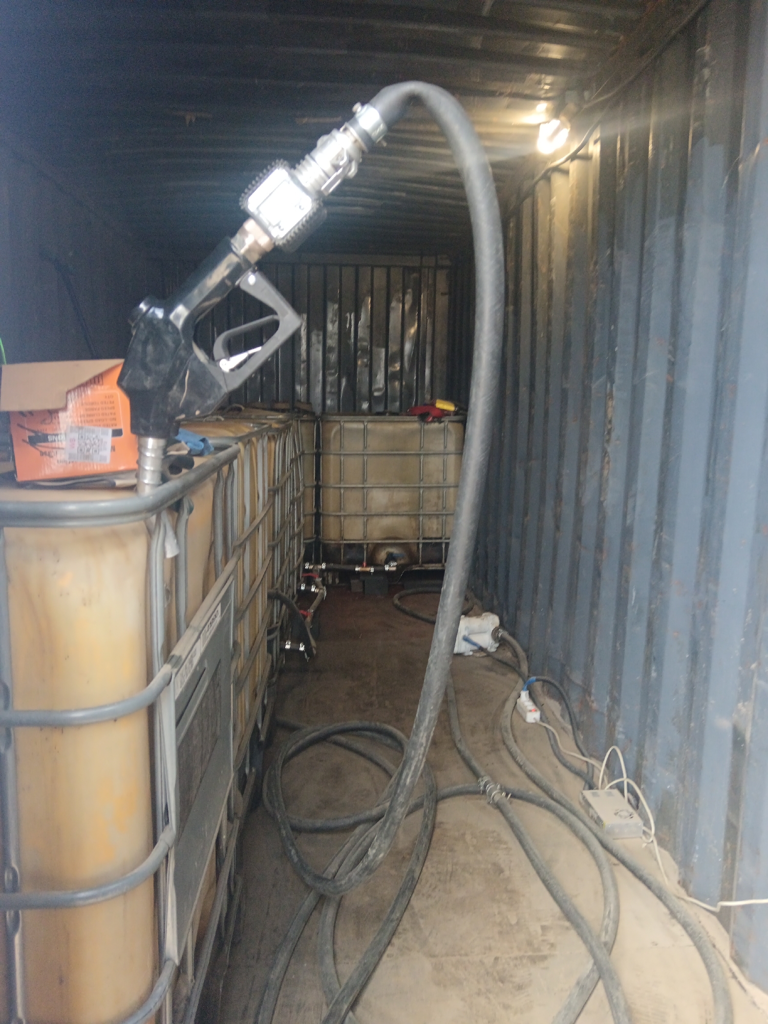 The image shows the interior of a shipping container in what appears to be an industrial setting. A row of large, caged, yellow/tan liquid totes lines the left side of the container, with a black fuel dispensing nozzle and hose positioned on top of the front totes. Cables, hoses, and electrical equipment are scattered across the floor. Other liquid totes are visible in the back of the container. The walls of the container are corrugated metal, lit by a single overhead light. The location is Яльгелево, Russia.