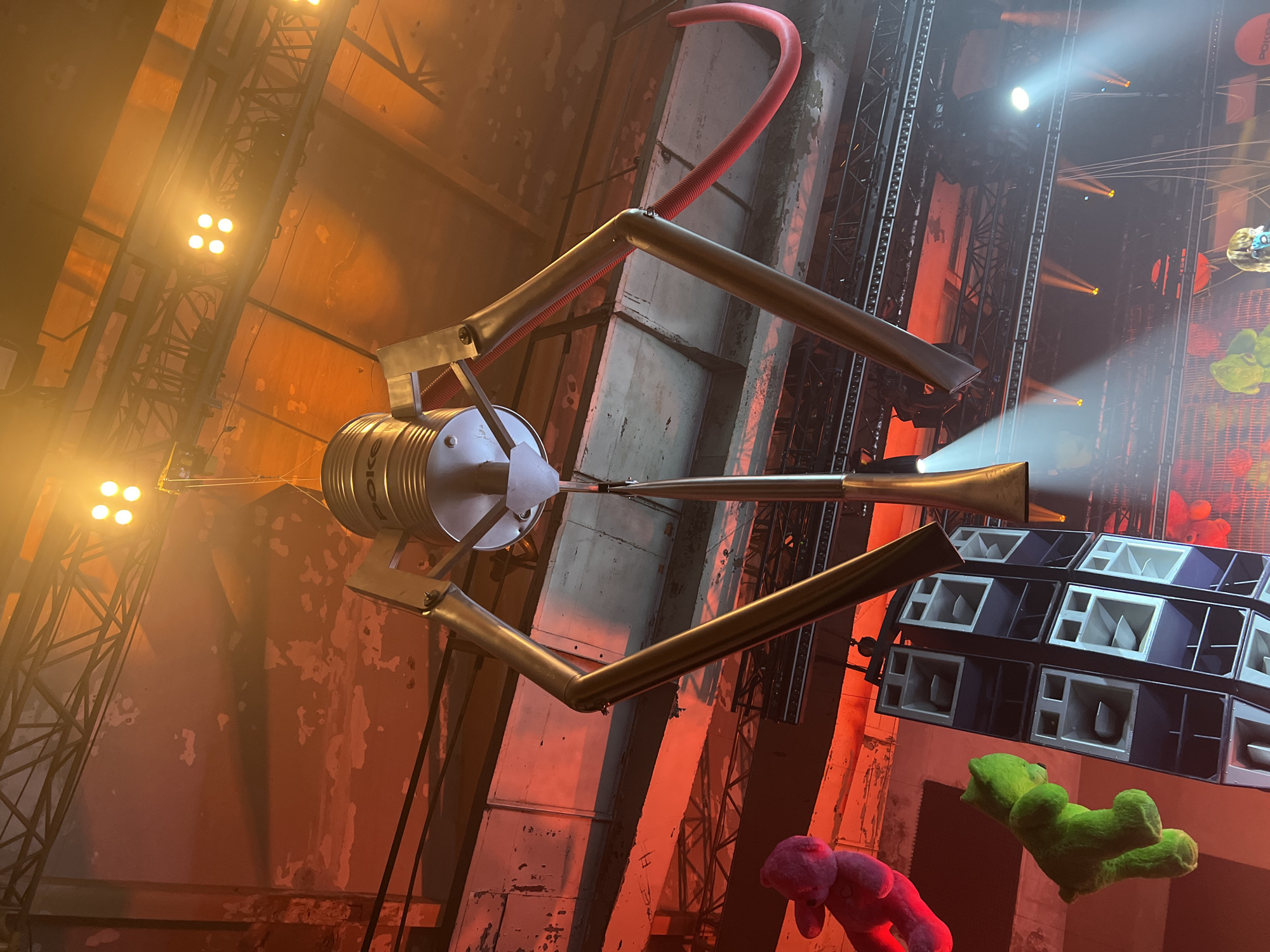This image depicts a large-scale artistic installation or stage setup within an industrial-style indoor venue, possibly a warehouse or concert hall. The central subject is an elaborate, metallic sculpture resembling a stylized insect, a satellite, or a musical instrument, suspended high above. It features a prominent cylindrical body with the text "POKEC" visible on its side, multiple long, jointed limbs, and two trumpet-like extensions that emit light. A curved red corrugated tube is also integrated into its upper structure.
The background reveals raw concrete walls with distressed surfaces and a complex network of steel truss systems, characteristic of industrial architecture adapted for events. Numerous professional stage lights are mounted on these trusses, casting intense yellow and orange illumination across the scene, creating a warm, hazy, and dramatic atmosphere with visible light beams.
On the right side of the frame, a large stack of professional sound speakers (a line array system) is clearly visible, suggesting a live music or performance event. Below the speakers, a vibrant green plush teddy bear is suspended, and a pink plush teddy bear is partially seen lower on the left, both appearing to float as part of the decorative elements. The perspective is upward-looking, highlighting the monumental scale of the installation and the venue's height. The scene suggests a dynamic, visually engaging event.