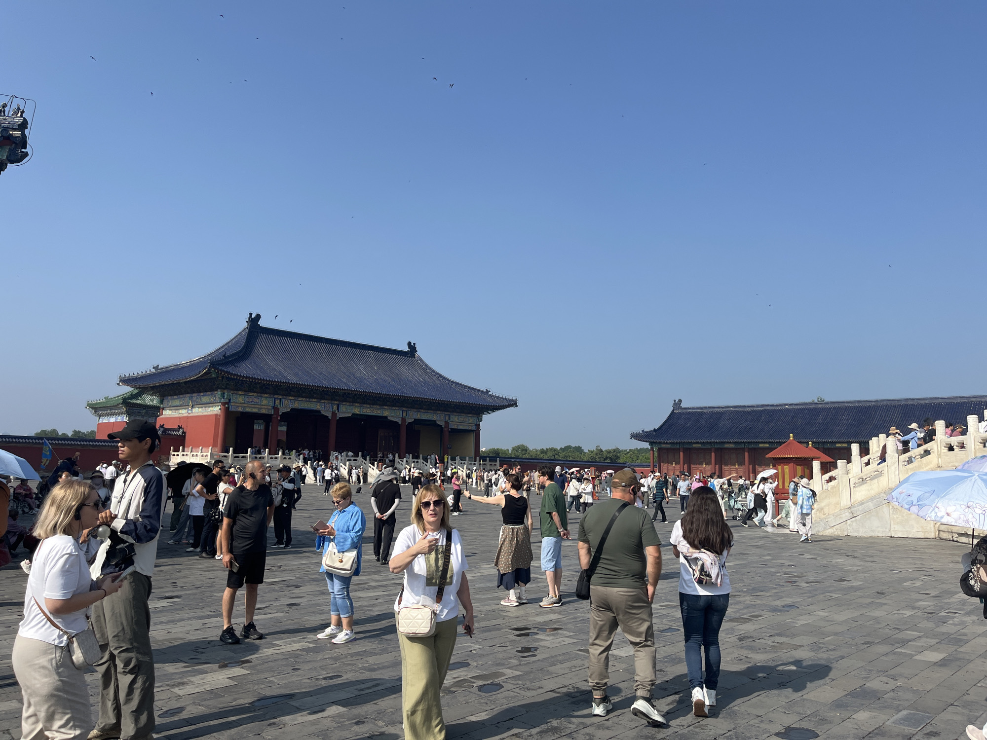 In an outdoor courtyard paved with grey flagstones, a group of tourists stands beneath a clear blue sky dotted with a few birds. Two traditional Chinese-style buildings with dark blue tiled roofs and red walls are visible in the background. The building on the left is larger and appears to be a hall, while the one on the right is smaller and has a red-roofed pavilion structure. Several people are walking around the courtyard, some holding umbrellas, and others are gathered in groups. In the foreground, a blonde woman wearing a white t-shirt and khaki pants is looking towards the camera and pointing. To her left, a man wearing a white and blue jacket and a black cap is standing. To the right of the woman, a man in a black t-shirt and shorts is looking at his phone, and a woman in a blue shirt and jeans is also looking at her phone. The overall scene is a sunny day at a historical site with many visitors.