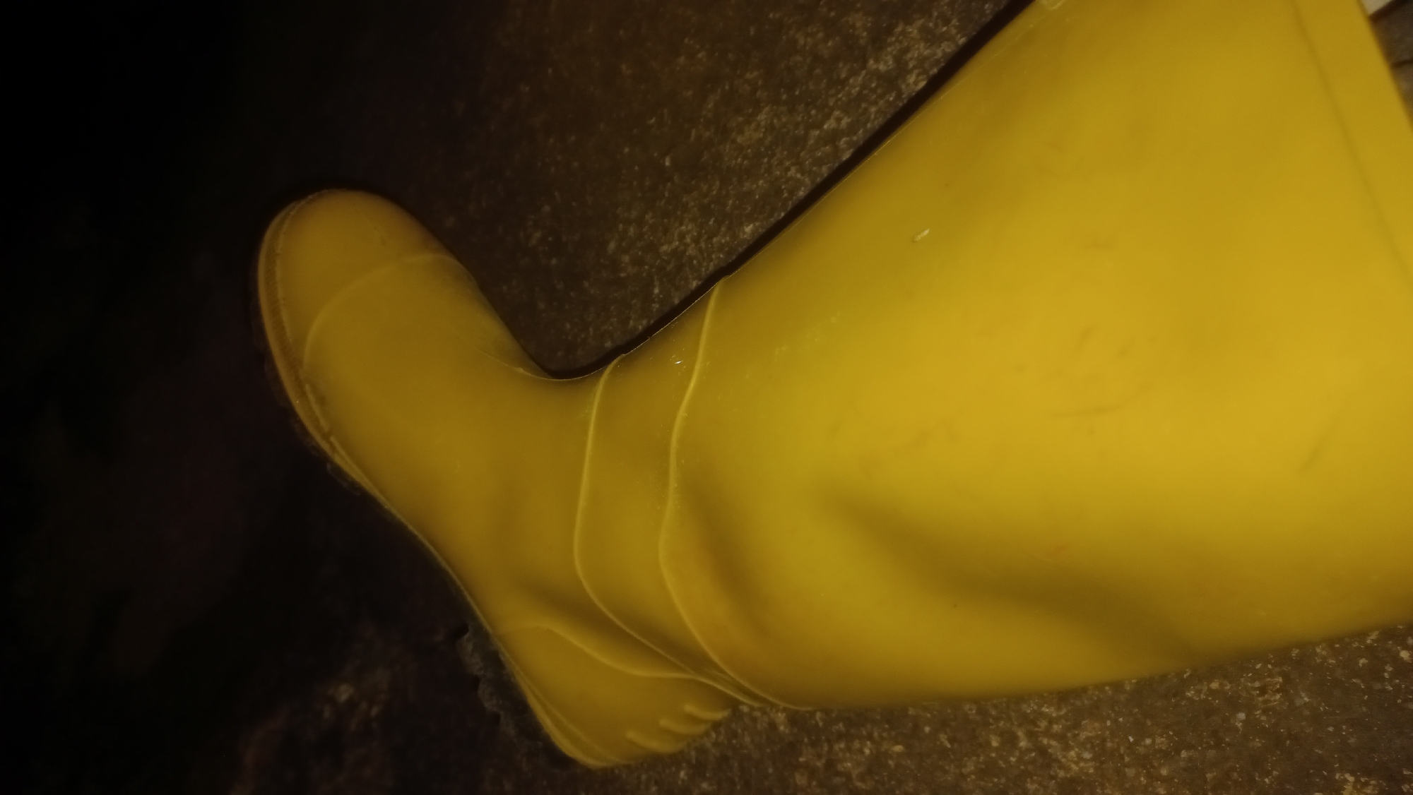 The image shows a close-up, low-angle shot of a bright yellow rubber boot. The boot is positioned diagonally across the frame, with its toe pointing towards the upper left and its top extending out of the frame to the right. The boot appears to be resting on a textured, dark-colored surface, possibly pavement or rough ground. The lighting is dim, casting shadows and highlighting the smooth, reflective surface of the rubber. The environment is dark, suggesting it might be nighttime or an indoor setting with low light. No people or specific landmarks in Kwang, Nigeria are visible. The image is a still photograph and does not depict any actions or interactions.