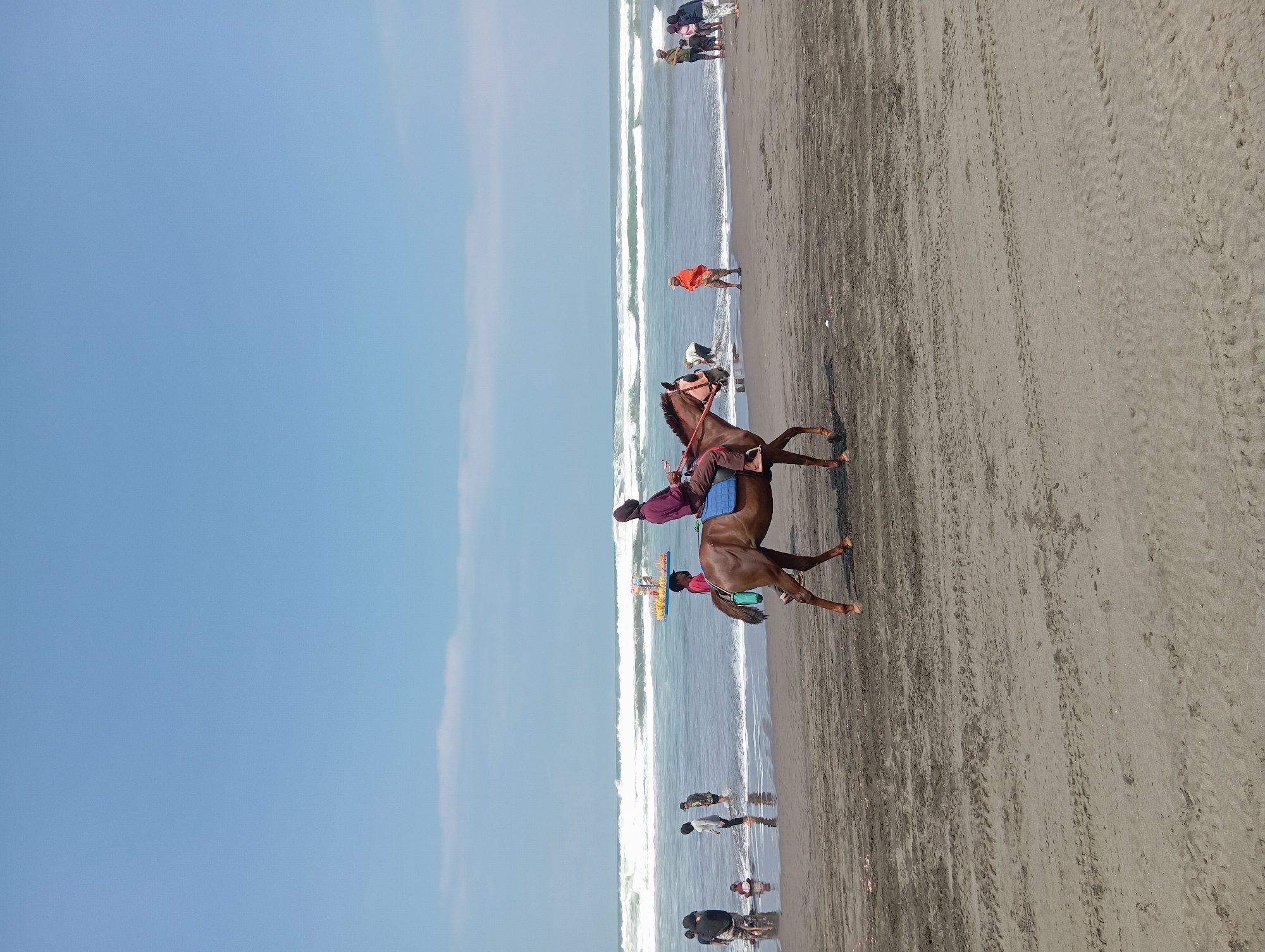 An overcast sky with a clear horizon hangs above a bustling beach scene in Parangtritis, Indonesia. In the foreground, a brown horse, equipped with a blue saddle, carries a rider adorned in a maroon shirt and headscarf. The horse's gait suggests a leisurely pace across the damp sand. The beach is populated with various individuals, some wading near the shore, others standing and observing the ocean. A small yellow boat with a canopy, possibly a vendor's stall, floats on the water. Waves crash onto the dark sand, creating a dynamic interplay between land and sea. The overall impression is one of a lively, communal gathering at the edge of the ocean.