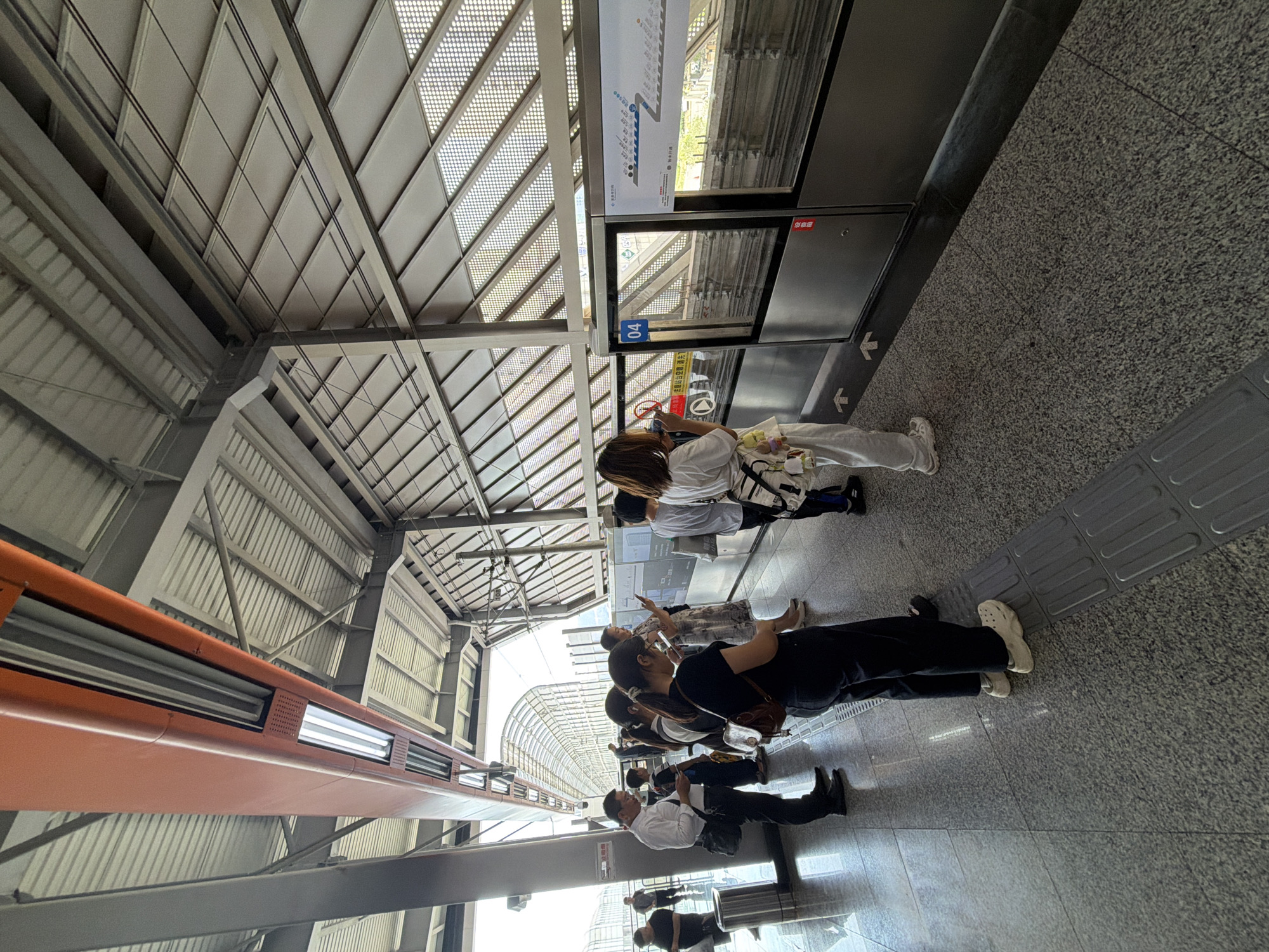 The image depicts a platform at the Tiaodeng station of the Shenzhen Metro in China, likely taken during daylight hours. Several individuals are standing on the platform, facing a set of glass doors that appear to be part of a station barrier. Most people are looking at their phones or towards the doors. One person in the foreground is wearing white Crocs. The platform has a grey, speckled floor with tactile paving for the visually impaired. Above the platform, a modern roof structure with corrugated metal and glass panels is visible, along with orange and grey support beams and industrial lighting fixtures. A digital display panel with route information is mounted on the glass barrier, indicating "04" which may refer to the train line or platform number. Outside the doors, a track and an empty train are partially visible. The overall environment suggests a well-maintained urban transit station.