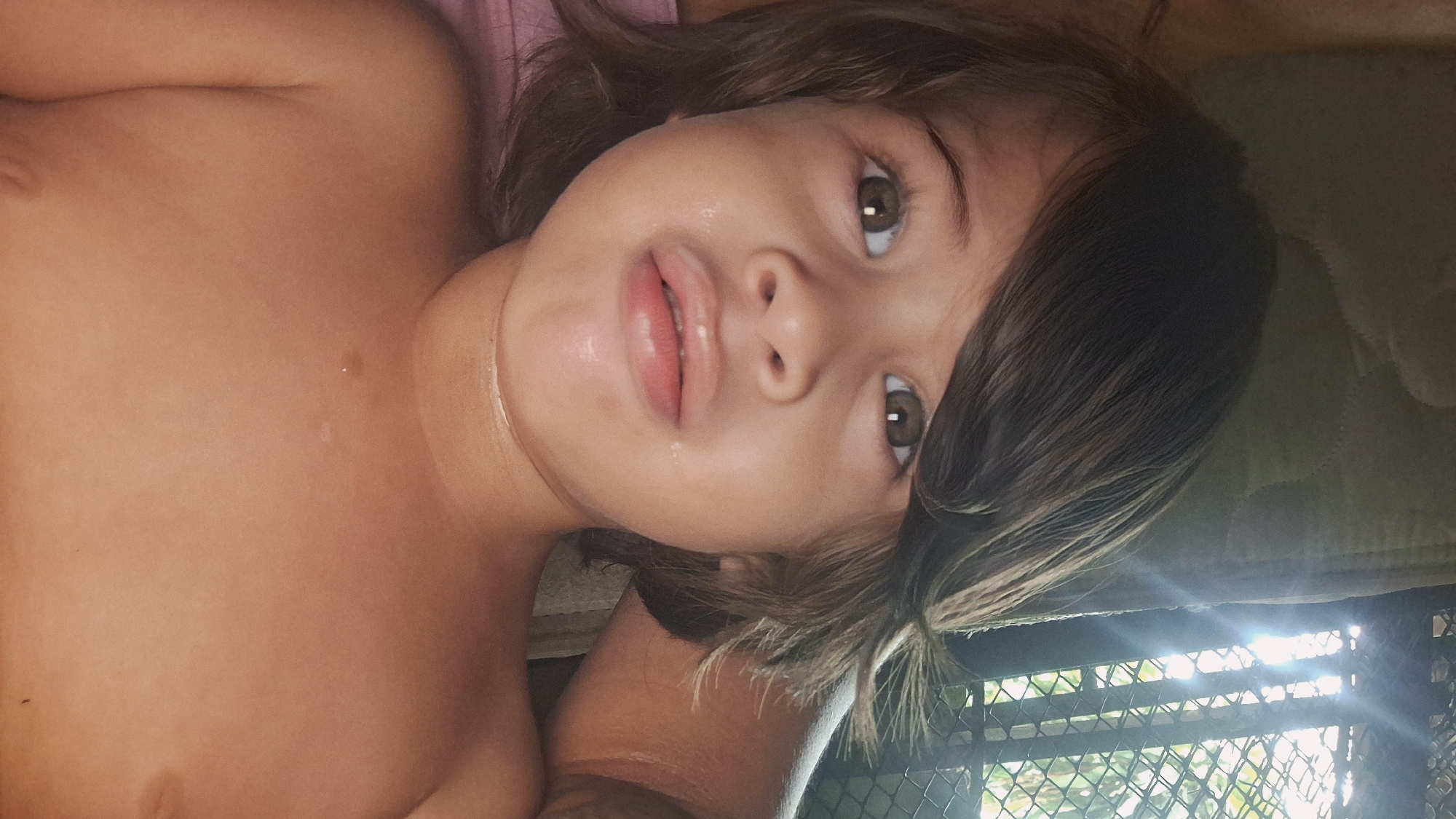 This is a close-up, horizontally oriented, undated, and likely personal photograph of a young child lying down. The child has brown hair, brown eyes, and fair skin. Their mouth is slightly open, and they appear to be looking towards the camera with a somewhat pouty or curious expression. The lighting suggests it might be daytime, with sunlight filtering in from the right side of the frame, creating a glare. The child is shirtless, revealing their chest and shoulder.
The background is dimly lit and appears to be an indoor or covered outdoor setting. To the right, a rough, textured surface, possibly a mat or cushion, is visible. Below this, a metal fence with a diamond pattern is present, through which a green, leafy outdoor environment can be seen, hinting at a tropical or subtropical location. The presence of sunlight and foliage suggests a warm climate.
Given the context "Vallecito, Venezuela," it's reasonable to infer the photograph was taken in this specific location. The image captures a candid moment, likely a snapshot taken by a parent or guardian. There is no visible text in the image.