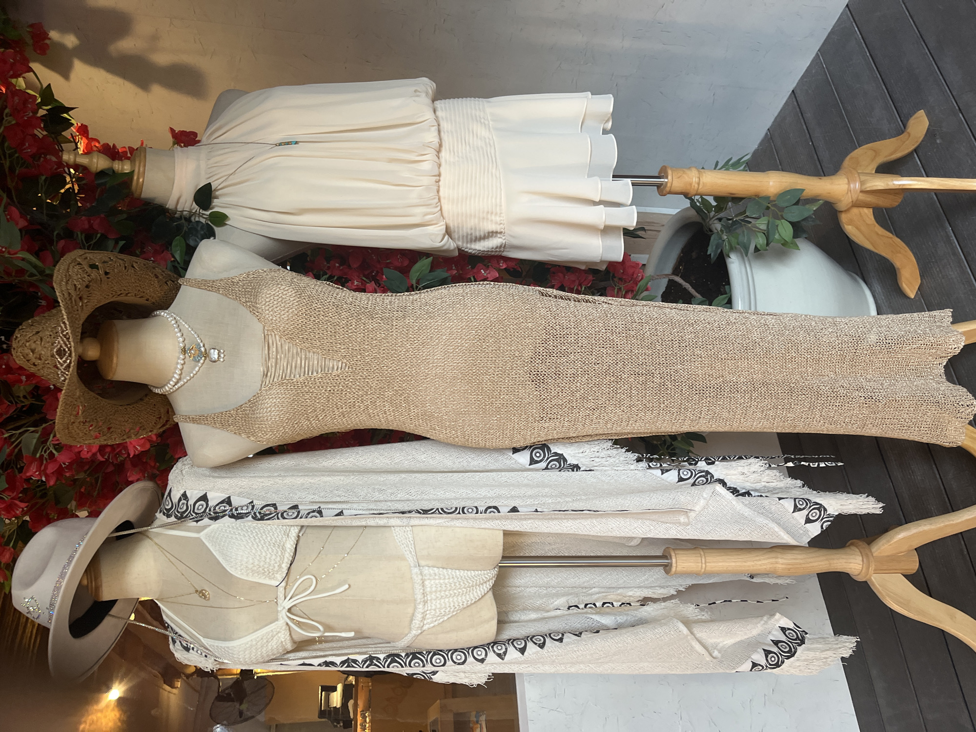 The image shows a retail display of women's clothing on three mannequins, set against a backdrop of red flowers and a textured wall. From left to right, the first mannequin is adorned with a white crocheted bikini, a long, sheer white kimono with black and white patterned trim and fringe, and a light grey wide-brimmed hat embellished with rhinestones. The second mannequin wears a tan, open-knit maxi dress and a straw hat. The third mannequin is dressed in a cream-colored, high-necked, flowing top with a cinched waist and a ruffled, tiered skirt. The display is situated outdoors, as indicated by the wooden floorboards and planters. This scene is not specifically identifiable as being located in Dubai, United Arab Emirates, and no details within the image allow for a definitive location within that city.