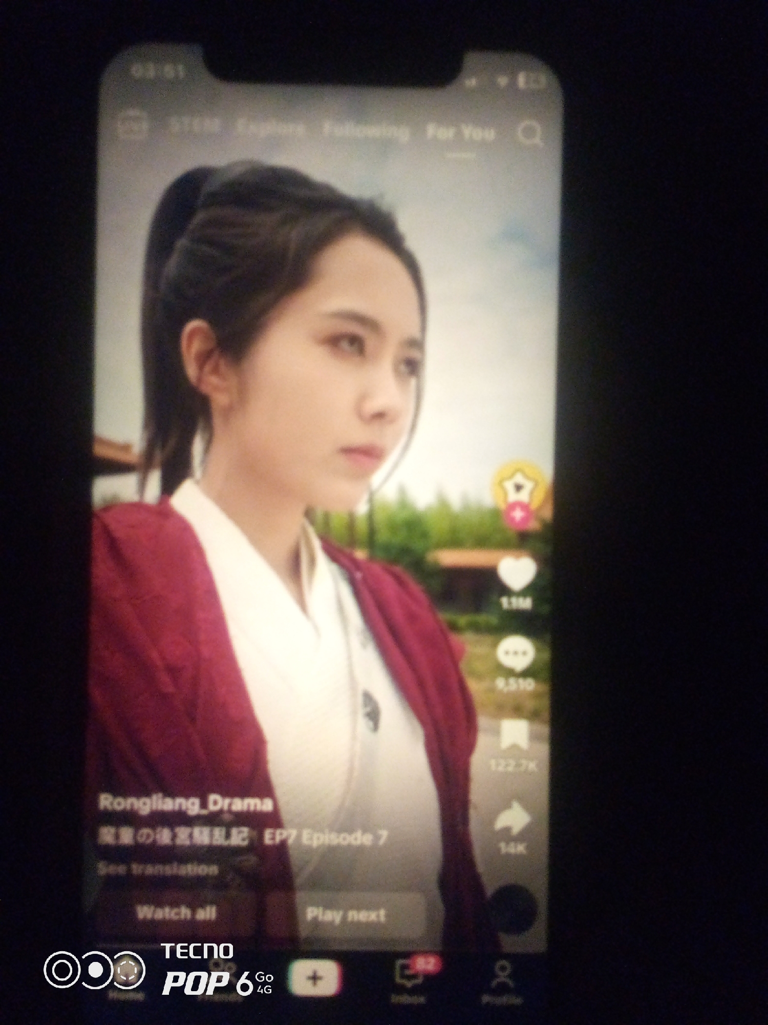 The image shows a close-up of a smartphone screen displaying a video. The video features a young East Asian woman with dark hair pulled back in a ponytail, wearing a maroon robe over a white garment. She is looking to her right, with a neutral expression. The background of the video appears to be an outdoor scene with greenery and some structures.
Overlaying the video are various interface elements typical of a social media app, likely TikTok. These include:
* **Top Bar:** Time "03:51" on the left, signal and battery indicators on the right. A navigation bar with "STEM," "Explore," "Following," "For You," and a search icon.
* **Right Side Icons:** A star icon with a plus sign, a heart icon with "1.1M" below it, a comment icon with "9,510" below it, a bookmark icon with "122.7K" below it, and a share icon with "14K" below it.
* **Bottom Left:** The text "Rongliang_Drama," followed by Chinese characters and "EP7 Episode 7." Below that, "See translation" and "Watch all" and "Play next" buttons.
* **Bottom Banner:** "TECNO POP 6 4G" branding with concentric circles and a plus icon. Additional icons resembling a home icon, a profile icon, and a notification icon are visible.
The image is a photograph taken of the smartphone screen, resulting in some glare and blurriness, particularly around the edges. The overall lighting suggests the photo was taken in a dimly lit environment. There is no information within the image that indicates the location is Howayi, Nigeria; this detail appears to be external information not derivable from the visual content itself.