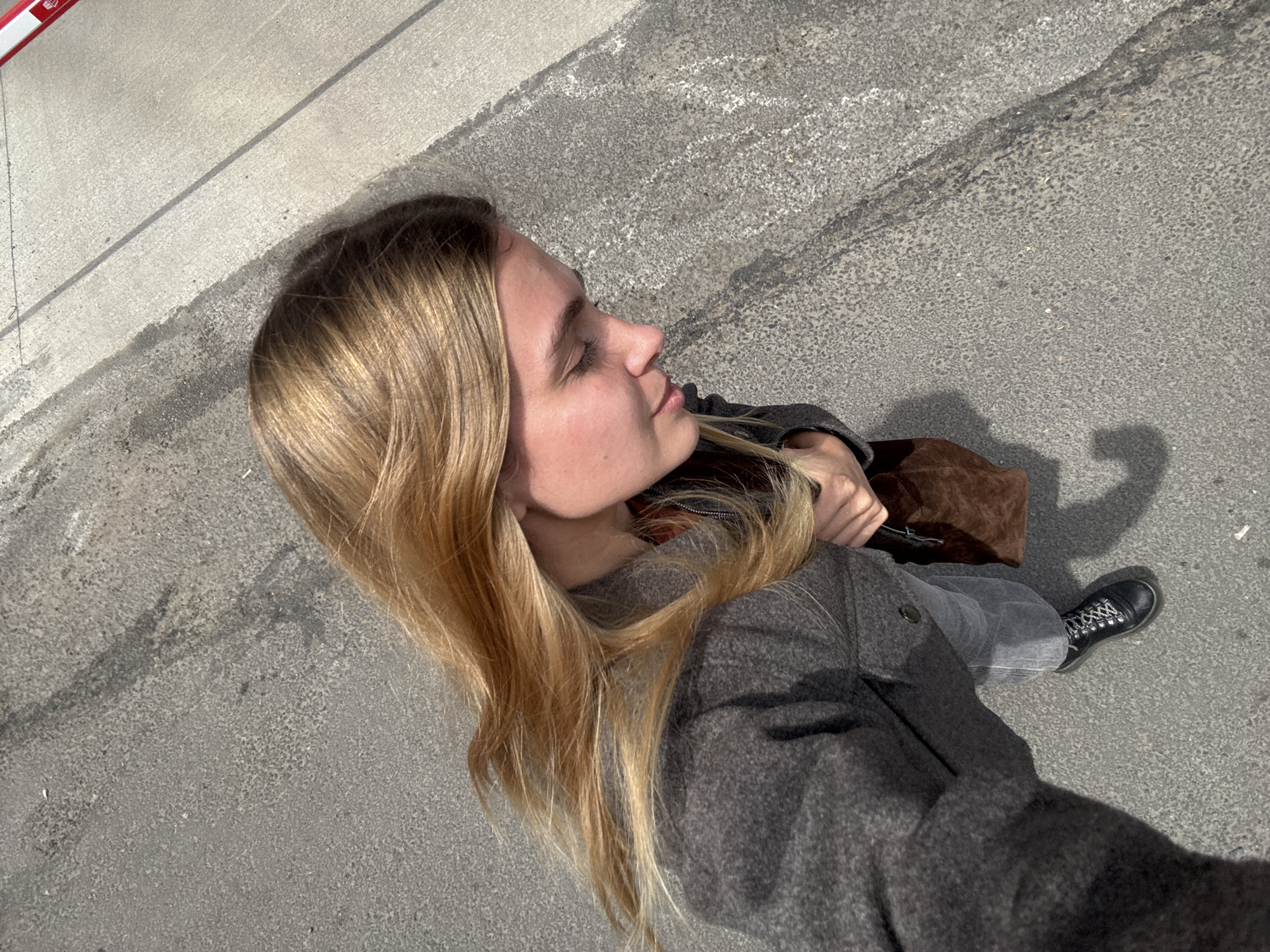 A young woman with long, blonde hair stands on a gray concrete surface. She wears a gray jacket, gray jeans, and black boots. She is carrying a brown bag. The location appears to be a city sidewalk.