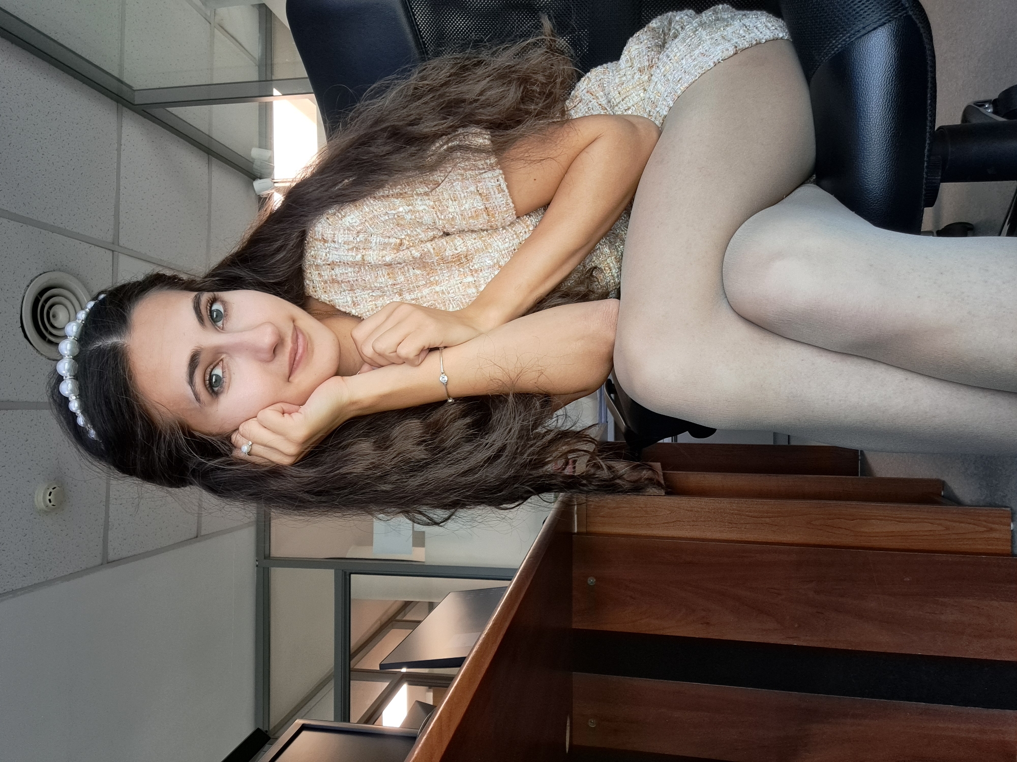 A young woman is sitting in an office chair with her legs crossed. She is wearing a light-colored tweed dress and light pantyhose. She has long, dark, wavy hair, a pearl headband, and is resting her head on her hand, smiling slightly at the camera. Her right arm is extended, and a bracelet is visible on her wrist. A ring is visible on her left ring finger. The office chair is black leather. The ceiling is white with a visible air vent. A portion of a wooden desk or shelving unit is visible in the foreground. The location is likely an office interior, but specific details to pinpoint the location in Sofia, Bulgaria are not discernible from the image.