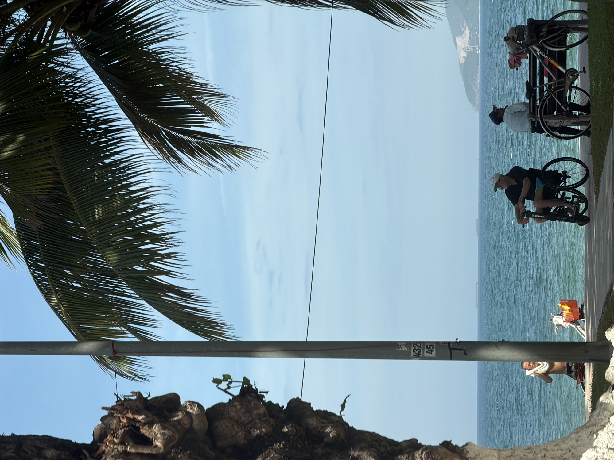 A group of people are enjoying a sunny day on the beach in Nha Trang Ward, Vietnam. In the foreground, a man in a wheelchair is talking to another man sitting on a bench next to a bicycle. Both men are looking out at the ocean. In the background, a third man is standing by the water, wiping his face with a towel. The ocean is calm, and there are a few ships in the distance. Palm trees are in the foreground, providing shade. A utility pole with numbers "432" and "45" is visible in the center of the frame. The scene suggests a relaxed and pleasant atmosphere.