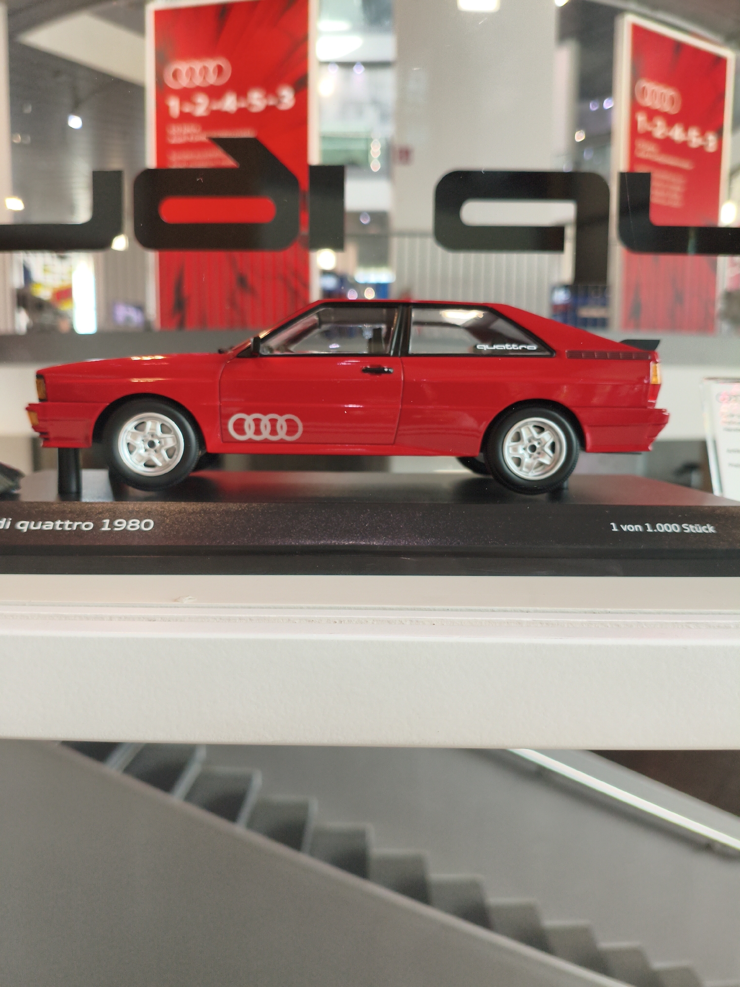 The image displays a red Audi Quattro model car positioned on a black display base. The car has white rims and the Audi logo is visible on its side. Text on the base reads "di quattro 1980" and "1 von 1.000 Stück". In the background, a large red poster with the Audi logo and numbers "1-2-4-5-3" is displayed, as well as other Audi branding. The overall setting appears to be an indoor exhibition or showroom, potentially at the Audi Forum in Ingolstadt, Germany, given the prominent Audi branding and the context of a car display. There are no people visible in the image.