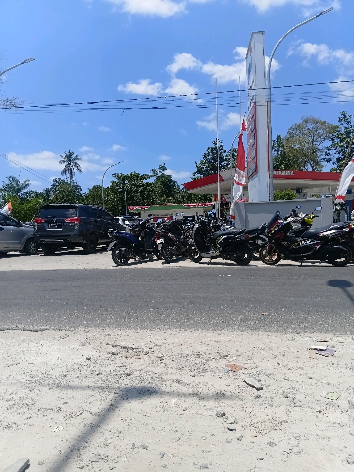 The image shows a street scene in Sungai Kapitan, Indonesia, on a bright, sunny day with a blue sky and scattered clouds. In the background, a Pertamina gas station with a tall white sign and red trim is visible. Several motorcycles are parked in front of the gas station. A dark-colored SUV is parked to the left of the motorcycles, and a silver car is partially visible on the far left. Trees and palm trees are scattered throughout the background, and utility lines are strung across the sky. The foreground consists of a paved road and a dirt shoulder with scattered debris. There are no people clearly visible in the image.