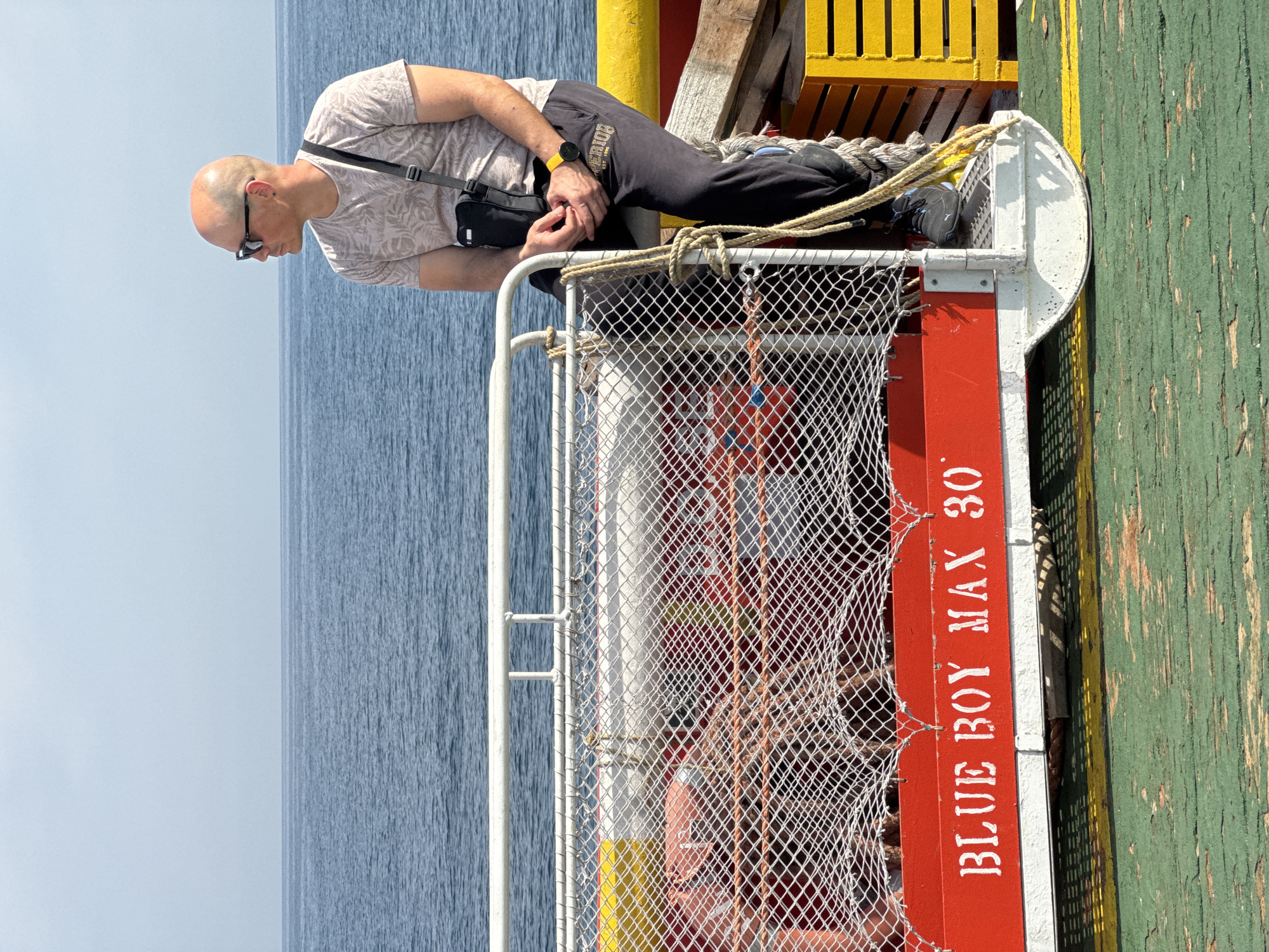 A bald, fair-skinned man wearing sunglasses, a light-colored short-sleeved t-shirt with a tropical pattern, dark sweatpants, and black athletic shoes is sitting on the edge of a boat or platform. He has a black crossbody bag slung over his right shoulder. He is looking out at the sea. The boat or platform has a red railing with white netting. The words "BLUE BOY MAX 30" are written in white on the red railing. The man's left hand rests on his knee, and his right hand is resting on his lap. He is wearing a black smartwatch on his left wrist. The scene is set in Marina di Ravenna, Italy. The sea is visible in the background. The platform is painted green and yellow.