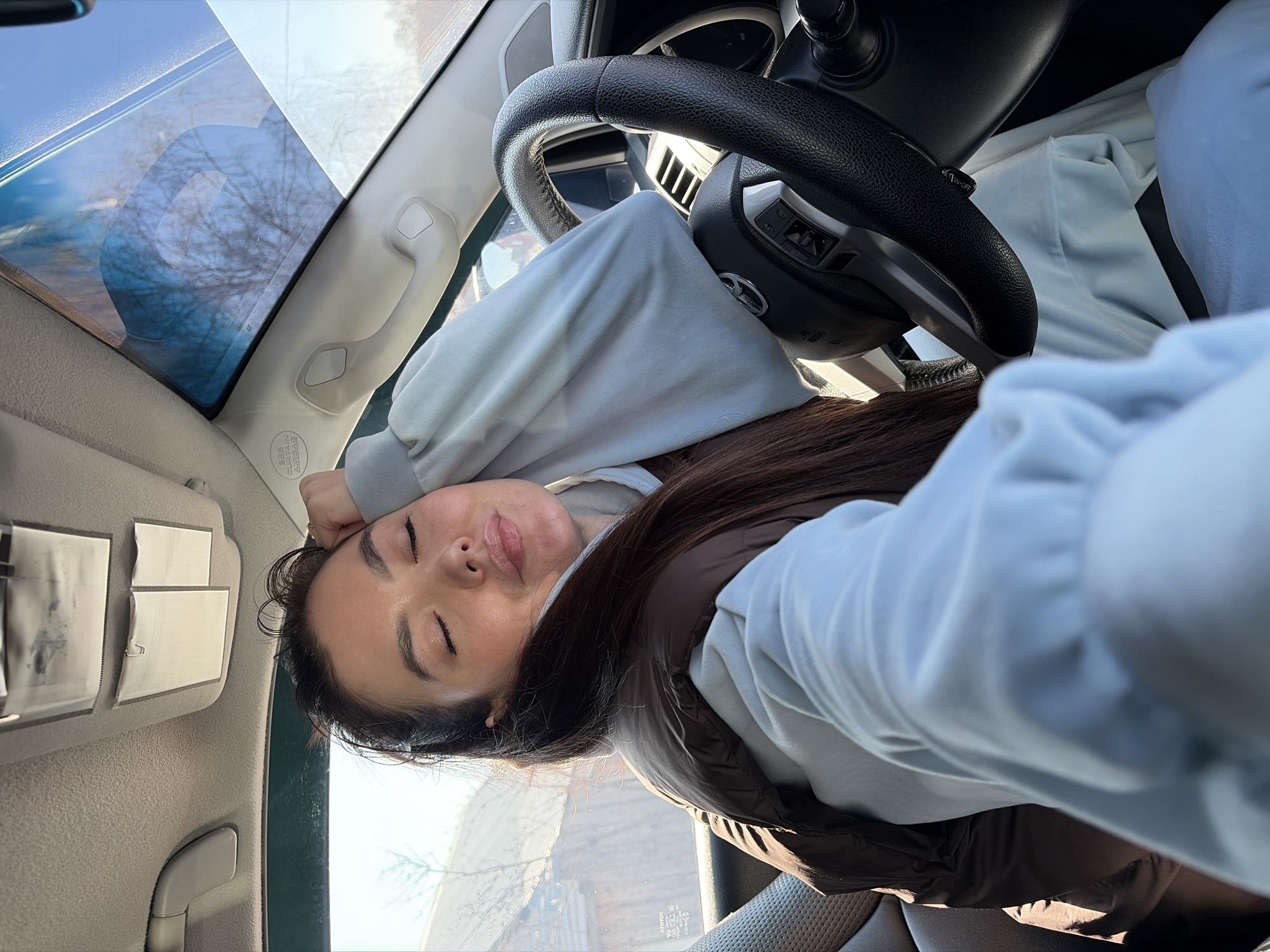 A woman with long brown hair, wearing a light blue hoodie and a brown puffer vest, is sitting in the driver's seat of a vehicle. Her eyes are closed and she appears to be resting her head on her hand, which is placed on the steering wheel. The interior of the car is visible, including the dashboard, windshield, and sun visor. Outside the windshield, trees and buildings are visible, giving context to a city location. While the city cannot be positively identified as Chaikovsky, Russia with the available information, this is what the request asks for.