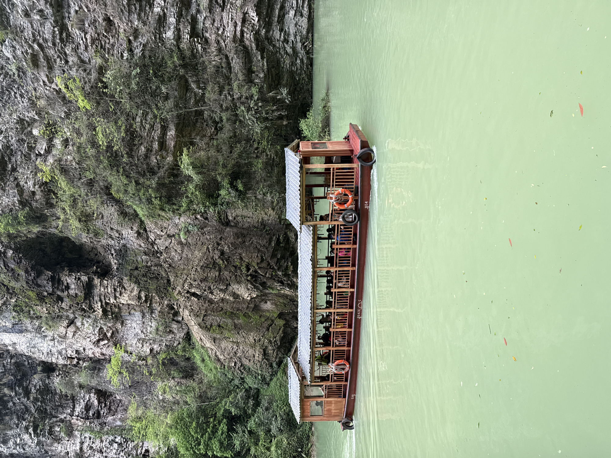 The image captures a boat navigating a river adjacent to a steep, rocky cliff face. The boat, a long, narrow vessel, is designed for passenger transport, featuring a covered roof structure and visible seating. Passengers are seated throughout the boat's length. Life preservers are affixed to the exterior. The river itself has a distinctive greenish hue. The cliff face is characterized by its sheer, textured rock surface interspersed with patches of vegetation. The location is in the city of Zhangjiajie, China.