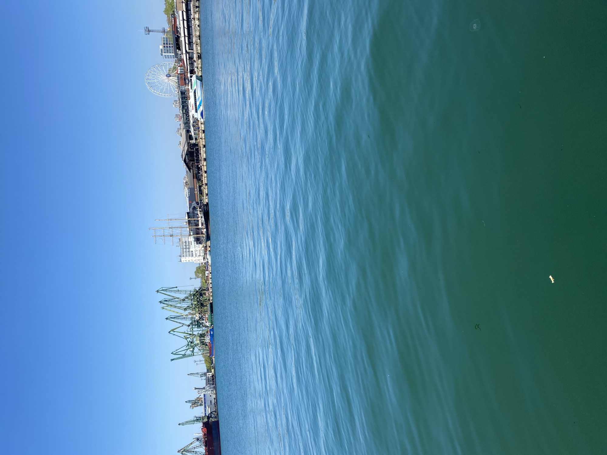 The image depicts a waterfront scene in Varna, Bulgaria. A portion of the coastline is visible, featuring cargo cranes, ships, buildings, and a Ferris wheel. The water is calm with mild ripples. The sky is a clear blue.
