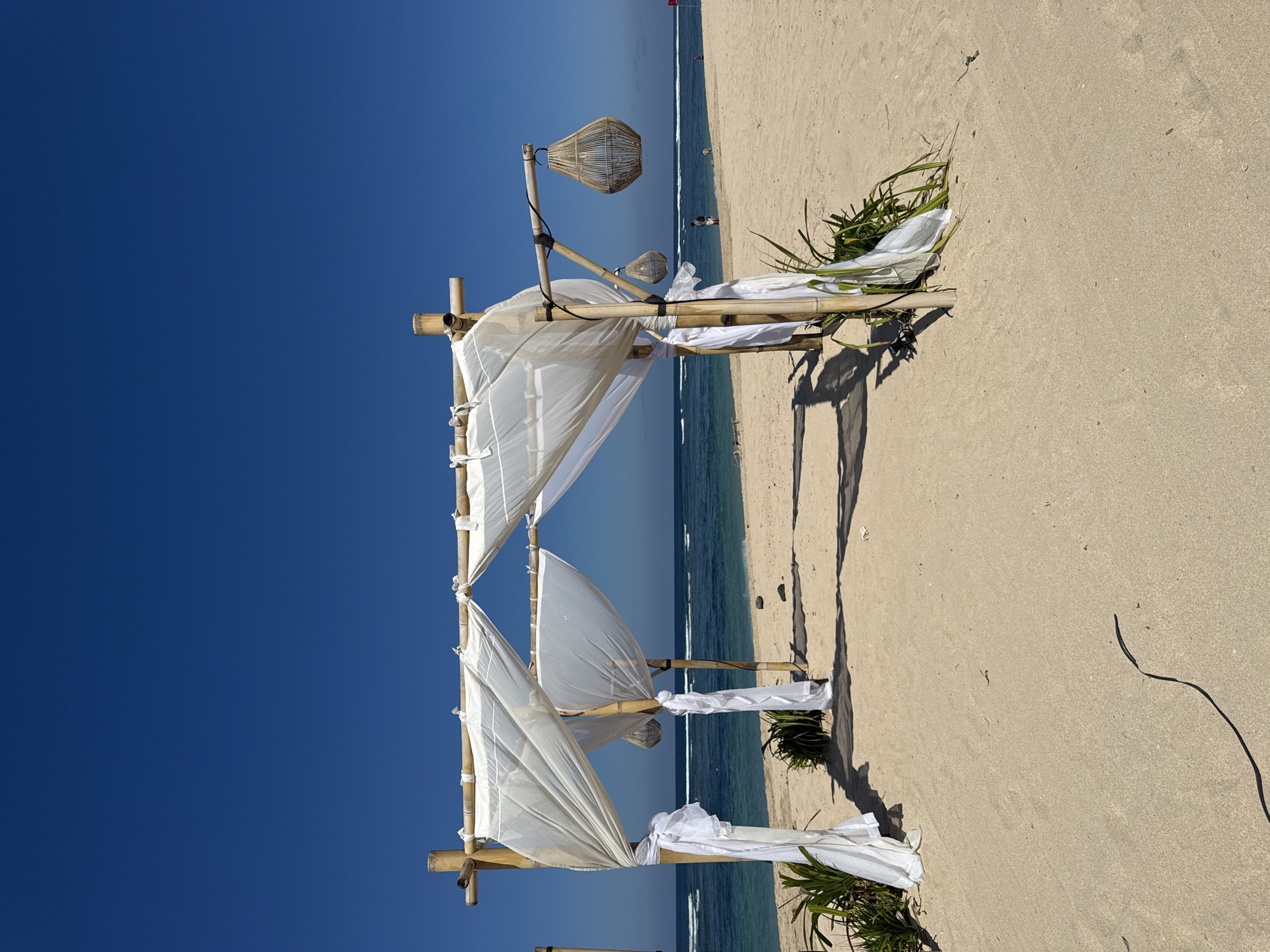 This is a wide-angle, eye-level shot of a bamboo structure with white fabric draped over it on a sandy beach. The structure appears to be set up for an event, possibly a wedding or ceremony. Two woven bamboo lanterns hang from the structure. In the background, the turquoise ocean meets a clear blue sky with gentle waves. Several small, dark objects are scattered on the beach. A few people are visible in the distance near the water's edge. The overall scene is serene and tropical, characteristic of locations such as Kuta Selatan, Indonesia, known for its beautiful beaches and resorts.
