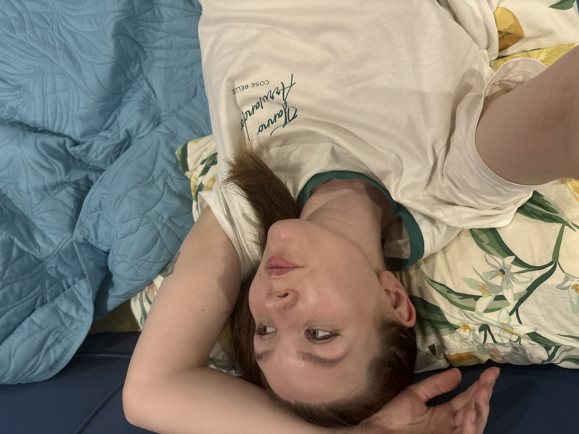 A woman lies on a bed with floral patterned sheets and a blue quilted blanket. She wears a white t-shirt with text in a dark font. She is indoors. It is not possible to confirm the location as Izhevsk, Russia.