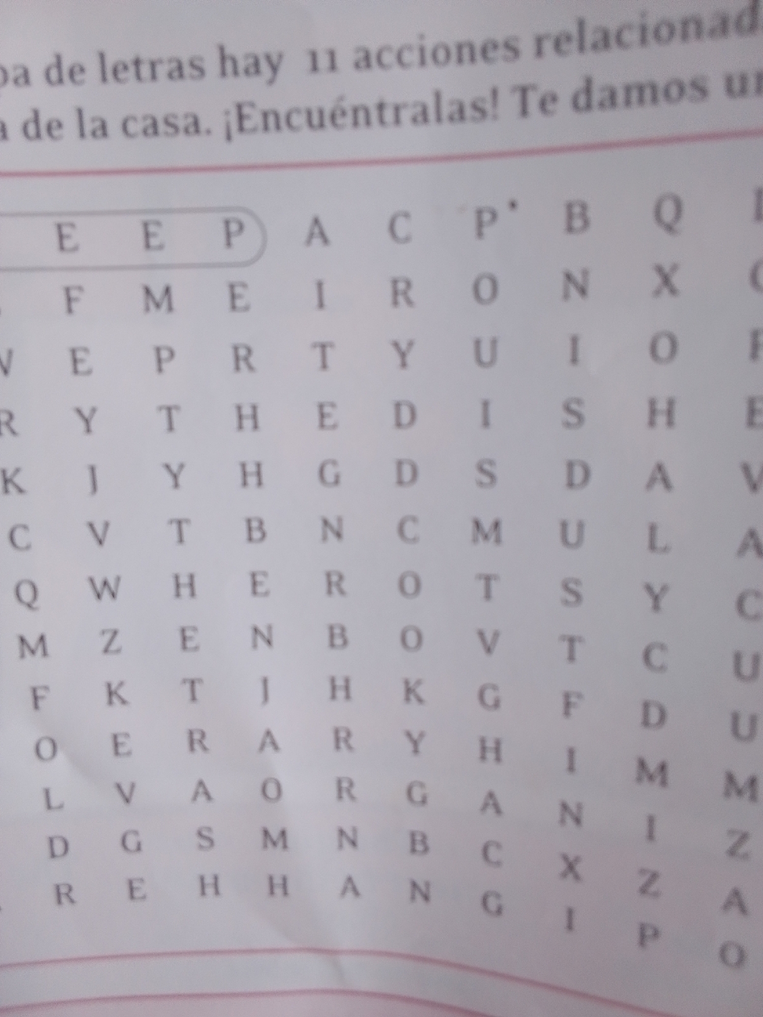 This media file is a close-up, slightly blurry photograph of a word search puzzle on a piece of paper. The puzzle appears to be in Spanish, with a title at the top that reads "Prueba de letras hay 11 acciones relacionadas con la casa. ¡Encuéntralas! Te damos un..." which translates to "Letter test there are 11 actions related to the house. Find them! We give you a...". The word search grid itself is filled with letters arranged in rows and columns. Some letters are circled or highlighted, suggesting they might be part of the solution or are being looked for. The background is plain white paper, and there are faint lines of a notebook or pad visible at the bottom of the image. The lighting is even, suggesting an indoor setting, likely daytime. No people or specific objects are depicted beyond the paper and the puzzle itself. The overall scene is that of an educational or recreational activity.