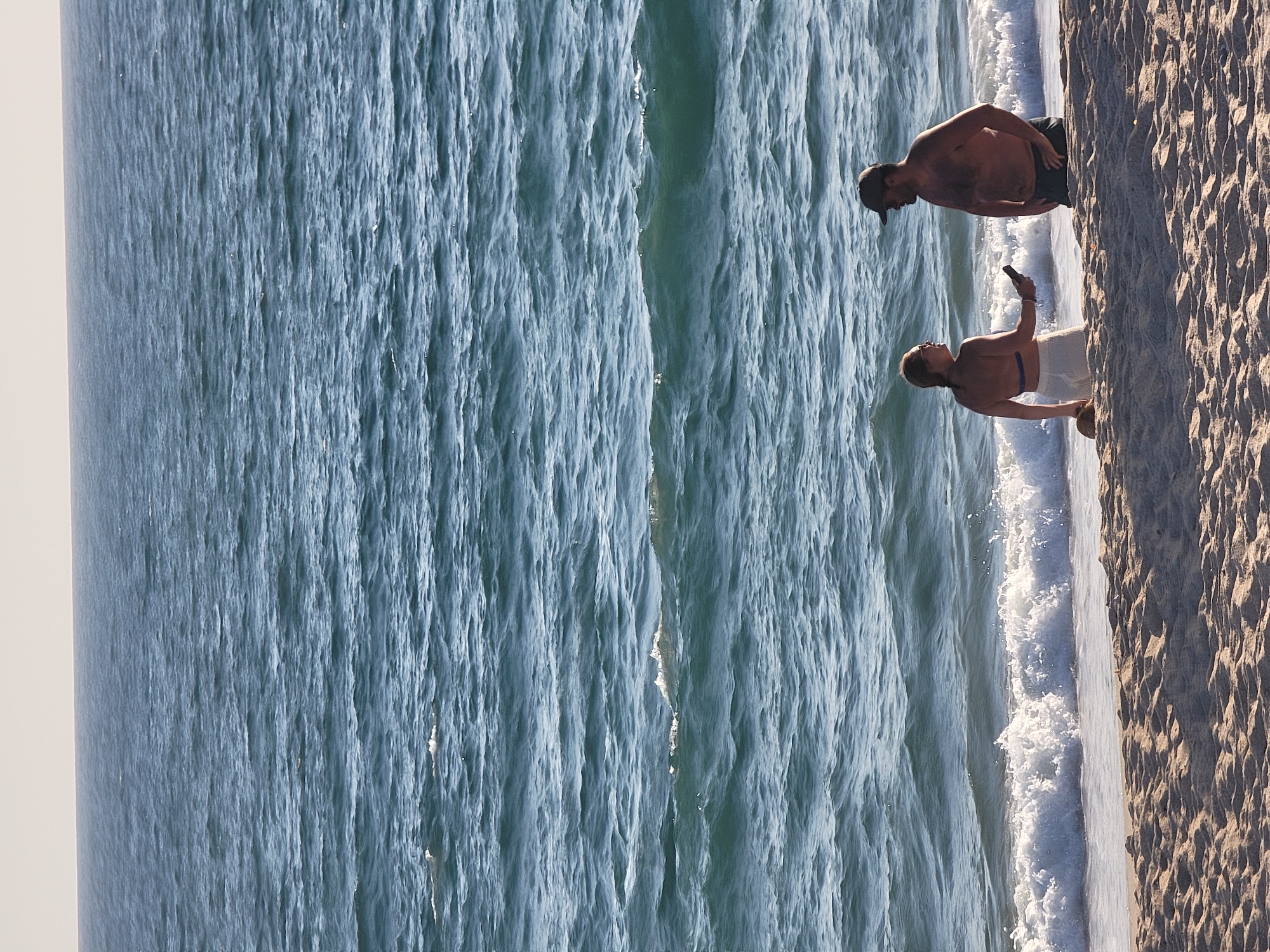 The image depicts a beach scene in Quarteira, Portugal, with two individuals standing near the water's edge. A woman, facing left, is wearing a blue bikini top and white shorts, holding a dark-colored object, possibly a phone. A man, facing the woman, is wearing a dark baseball cap and appears to be shirtless. Both are partially submerged in the ocean. The water is blue with moderate waves and white foam near the shore. The sandy beach is in the foreground, with ripples from the sand. The horizon line between the ocean and the sky is visible in the background.
