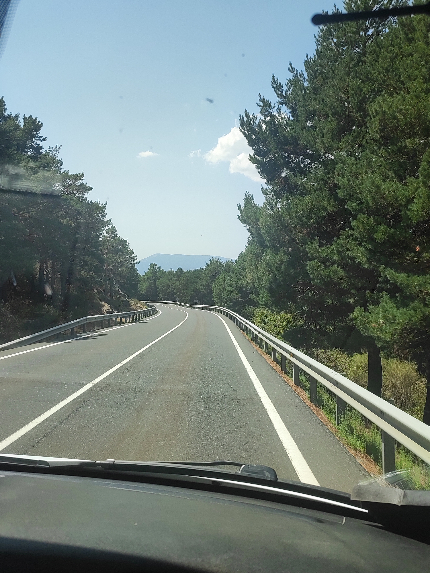 The image shows a view from inside a vehicle, looking out through the windshield at a winding road in a forested mountainous area. The road is asphalt with white lines marking the lanes. On the right side of the road, there is a metal guardrail. Tall pine trees line both sides of the road. In the distance, there are more trees and a hazy mountain range under a light blue sky with a few scattered clouds. The dashboard of the vehicle is visible at the bottom of the image. The location is the city of Navacerrada, Spain.