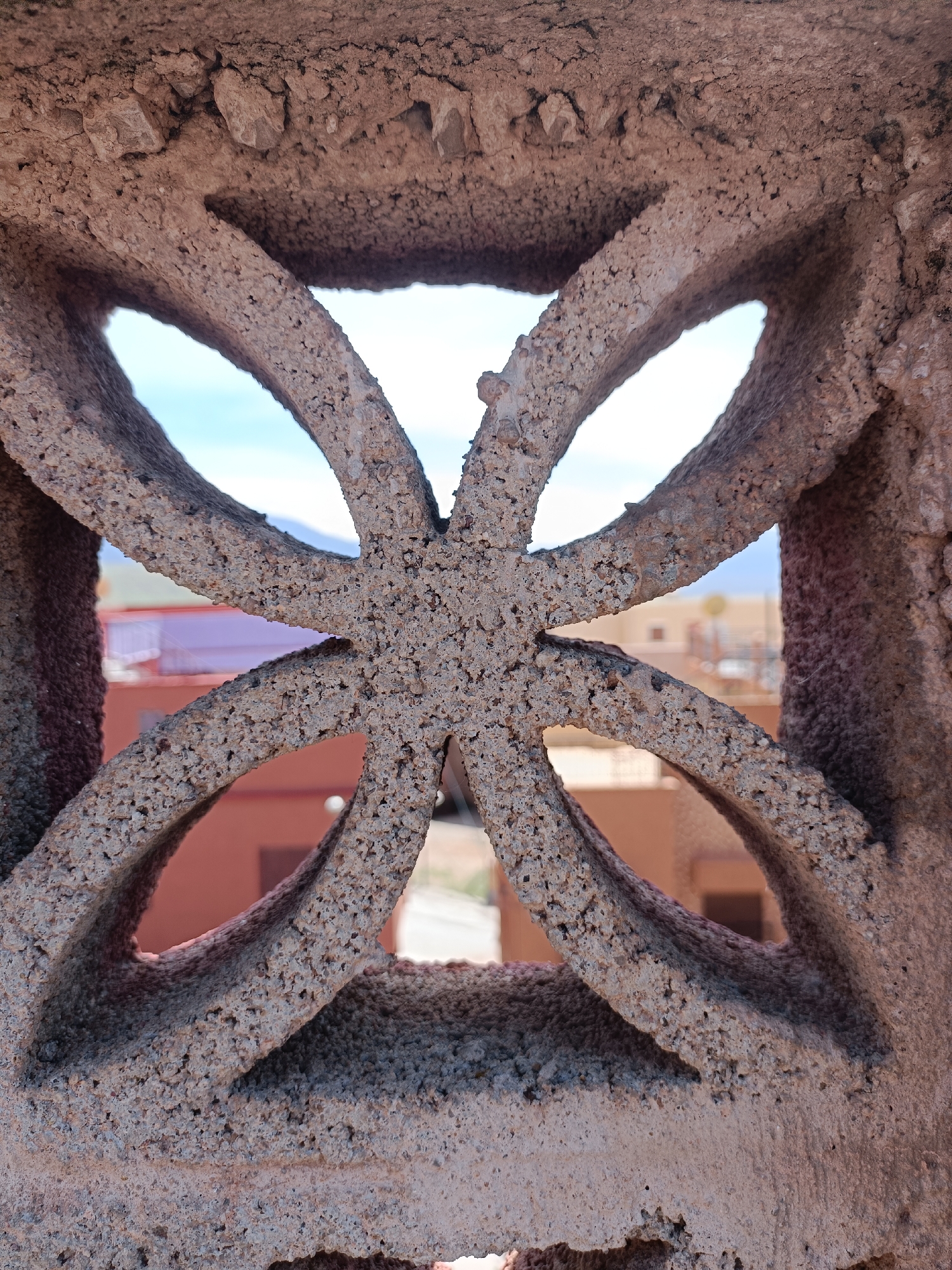 Close-up view of a decorative stone block featuring a symmetrical, clover-like design with open spaces between the curves. The rough, textured surface of the stone is a mottled tan color. Through the openings, a blurry urban landscape is visible, including rooftops, a building with a red roof, and a distant, hazy sky with a hint of blue and mountains. The location is Ouaouizert, Morocco, inferred from the architectural style.