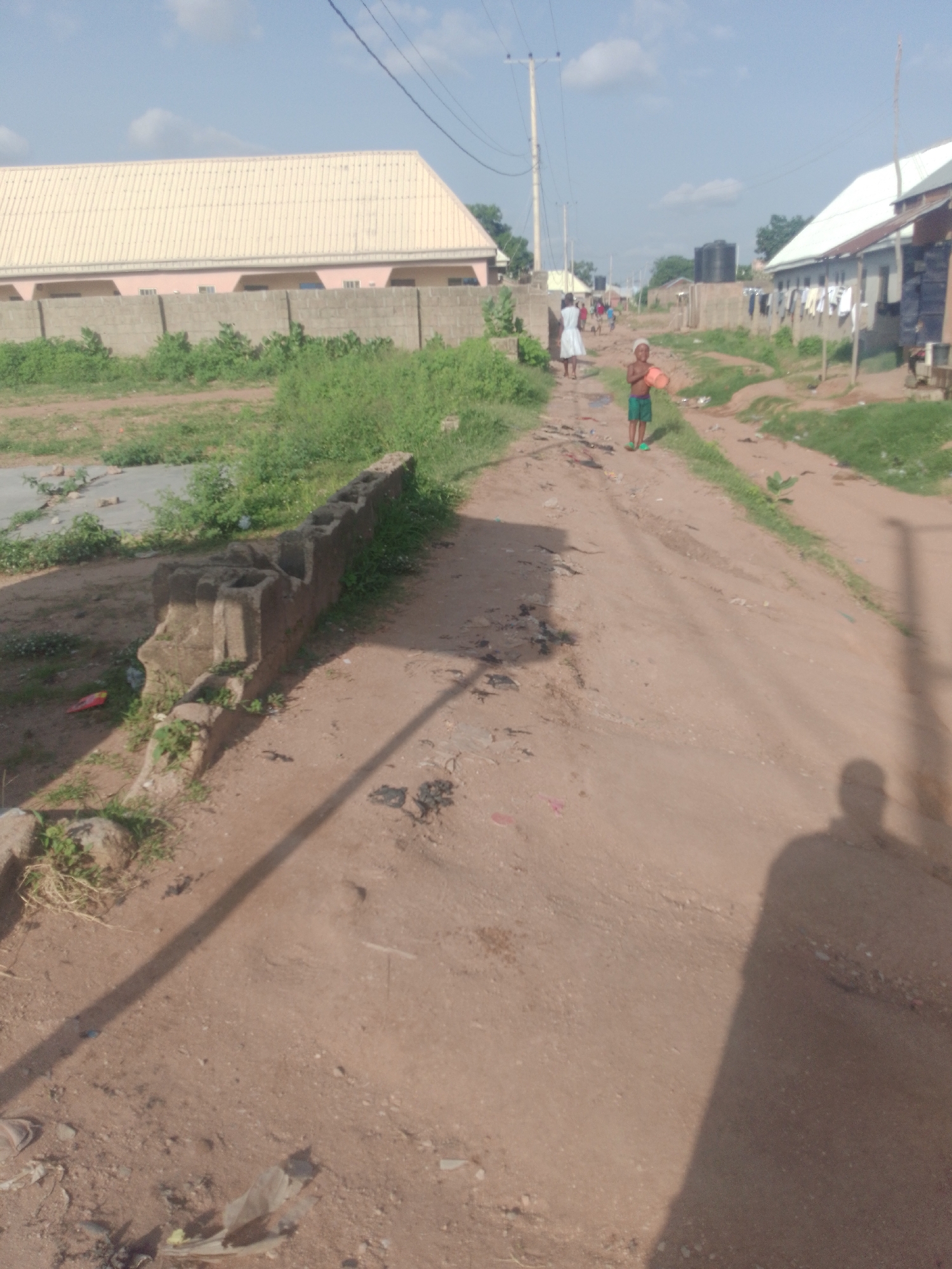 In Jalingo, Nigeria, an outdoor scene unfolds with a dirt road dominating the foreground and midground. On the right side of the road, a shadow of a person is cast across the dusty surface, stretching from the lower right corner upwards. A child, wearing green shorts and a light-colored top, stands in the middle of the road, holding a reddish, round object. Further down the road, a person in a white dress walks away from the camera.
To the left of the road, a low concrete barrier runs along the edge of a grassy area. Behind this barrier, a light-colored building with a corrugated metal roof and a pink facade is visible, partially obscured by greenery. Across the road, on the right side, other buildings with corrugated metal roofs line the street, with laundry hanging on lines between them. Water tanks are also present in the background. The sky above is a bright blue with scattered white clouds. The overall impression is of a residential area with unpaved roads and modest structures.