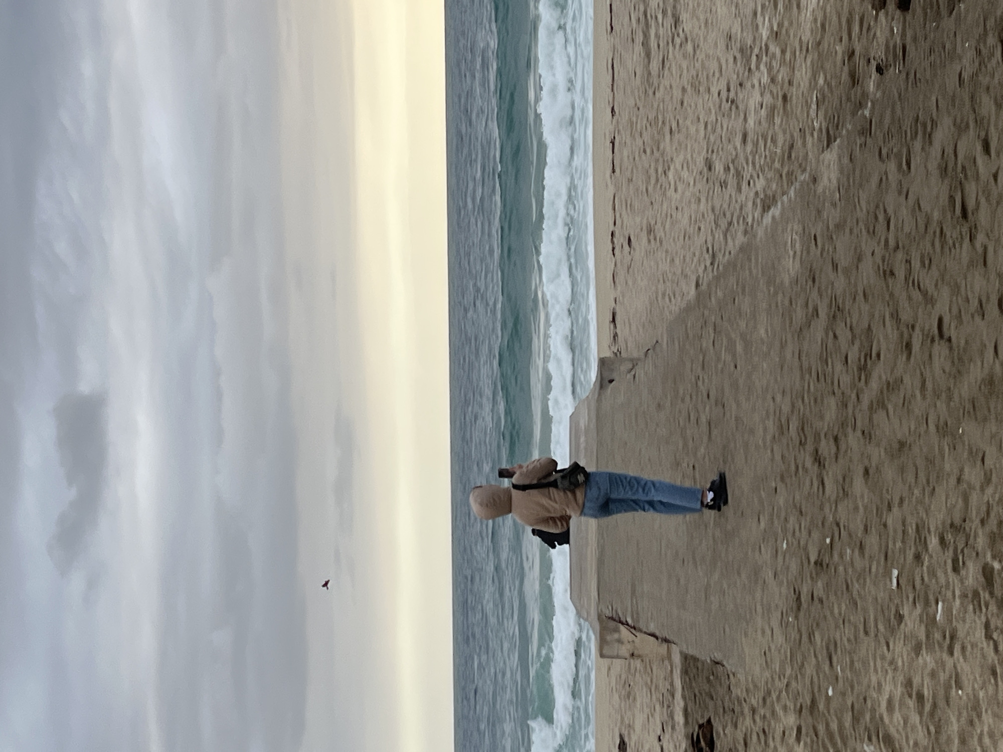 A person stands on a concrete structure, possibly a pier or breakwater, next to the ocean. The person is wearing a tan hooded jacket and blue jeans, and dark footwear. They appear to be looking at or photographing the sea, perhaps at or near the coast of Alanya, Turkey, judging by the cloudy sky and the presence of waves. A small, dark object resembling a bird is visible in the sky. The scene is dominated by the concrete, the ocean with white waves, and a cloudy sky with diffused light. The photograph is taken from a low angle or perspective, with the concrete structure taking up a large portion of the frame. The land is visible to the right of the concrete structure and is darker in comparison to the concrete structure.