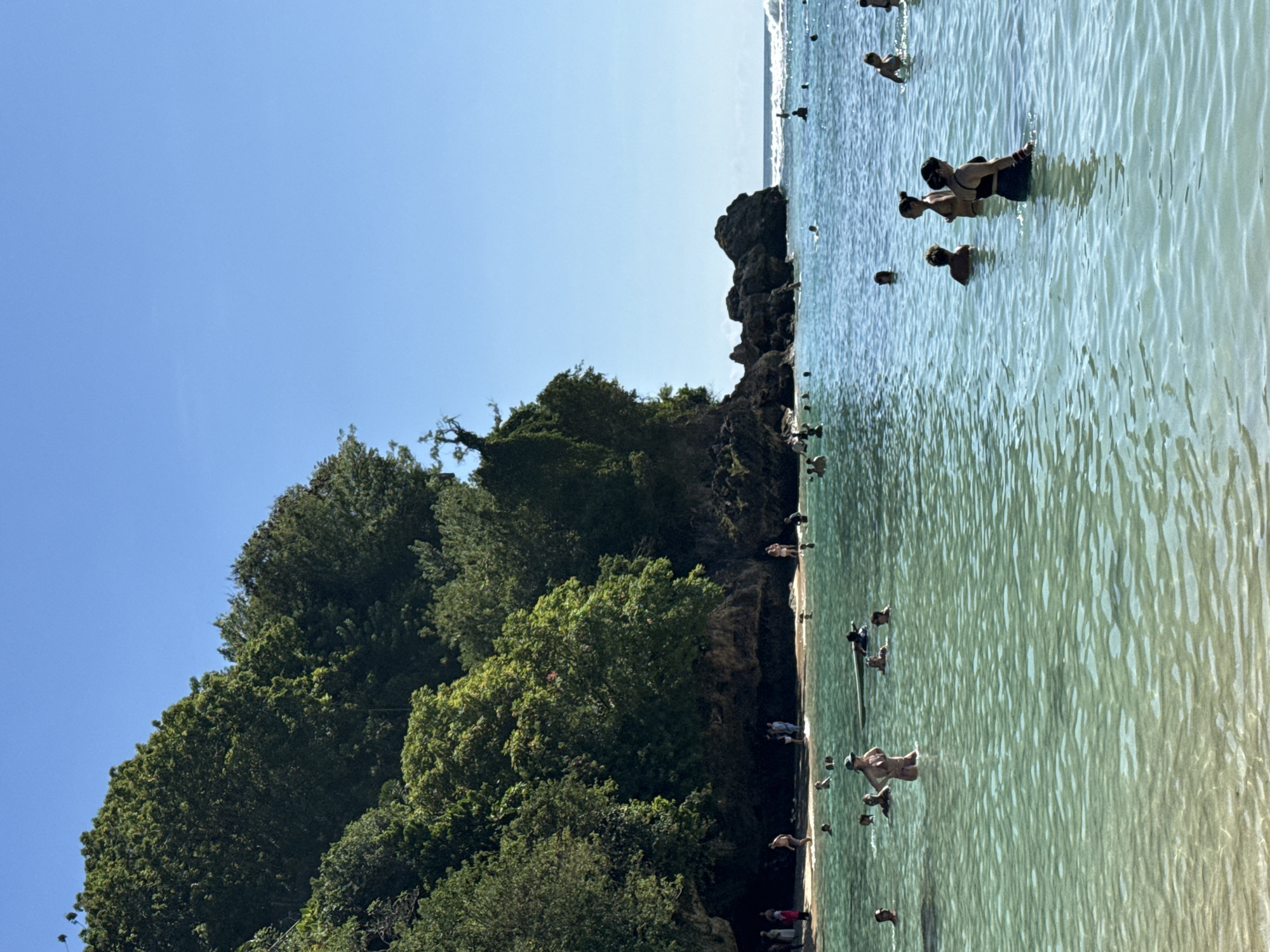 The image depicts a beach scene in Kuta Selatan, Indonesia, with numerous people in the turquoise water. Some are swimming while others appear to be standing in the shallows. The background features lush green vegetation on a rocky cliff that borders the shoreline. The sky is clear and blue.