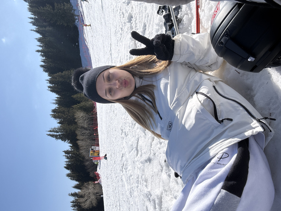 A young female is photographed reclining on a snow-covered ski slope under a clear blue sky in Bistritsa, Bulgaria. She has light brown hair, wears a black knitted beanie with a black pom-pom, a white ski jacket featuring a "L1K3" logo on the left chest, and white sweatpants with partial "PURE PROJECT" text visible on the left leg. Her lips are pursed into a kiss shape, and her left hand, covered by a black textured glove, displays a peace sign. To her right, a black ski helmet or equipment carrier and a red ski are partially visible on the snow. The background consists of a vast snowfield, a dense forest of evergreen trees, and red safety netting along the slope. A small yellow structure with a shovel and poles stands in the mid-ground left, and distant figures of skiers are present further up the incline.