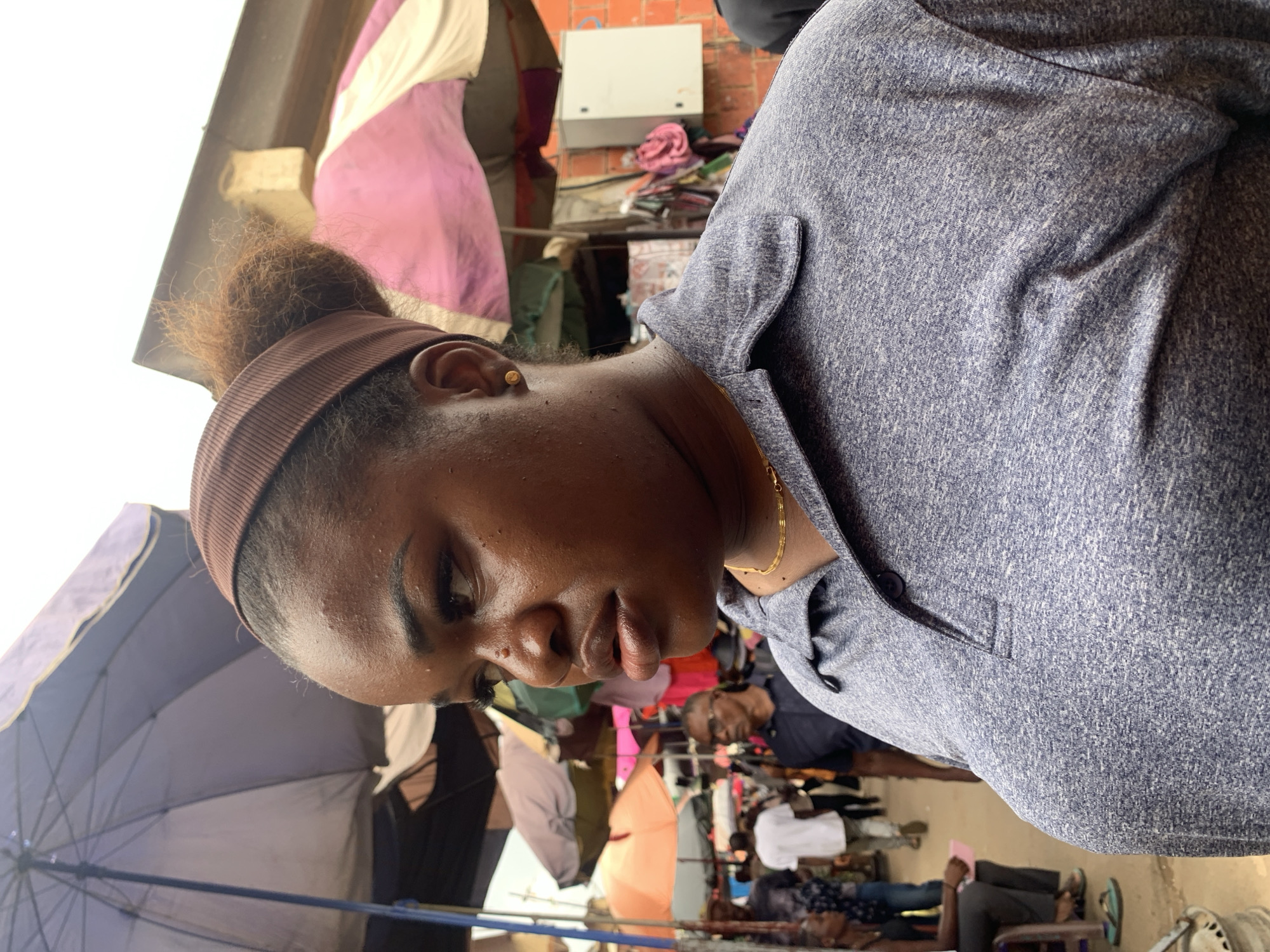This media file shows a close-up, vertically oriented, low-angle shot of a young Black woman. She is wearing a brown headband and a blue-grey heathered long-sleeved shirt with a small pocket and a button closure near her collarbone. A thin gold chain is visible around her neck. Her expression is pensive, with her eyes looking downwards. The background is blurred and shows an outdoor market scene. Various colorful umbrellas are visible, suggesting it might be sunny or a place with vendors using umbrellas for shade. In the distance, there are hints of brickwork, possibly buildings, and some racks of clothing or textiles. Several other people are visible in the background, some appearing to be shoppers or vendors. The overall impression is of a bustling marketplace environment in Abuja, Nigeria. The lighting suggests it is daytime, possibly with bright ambient light.