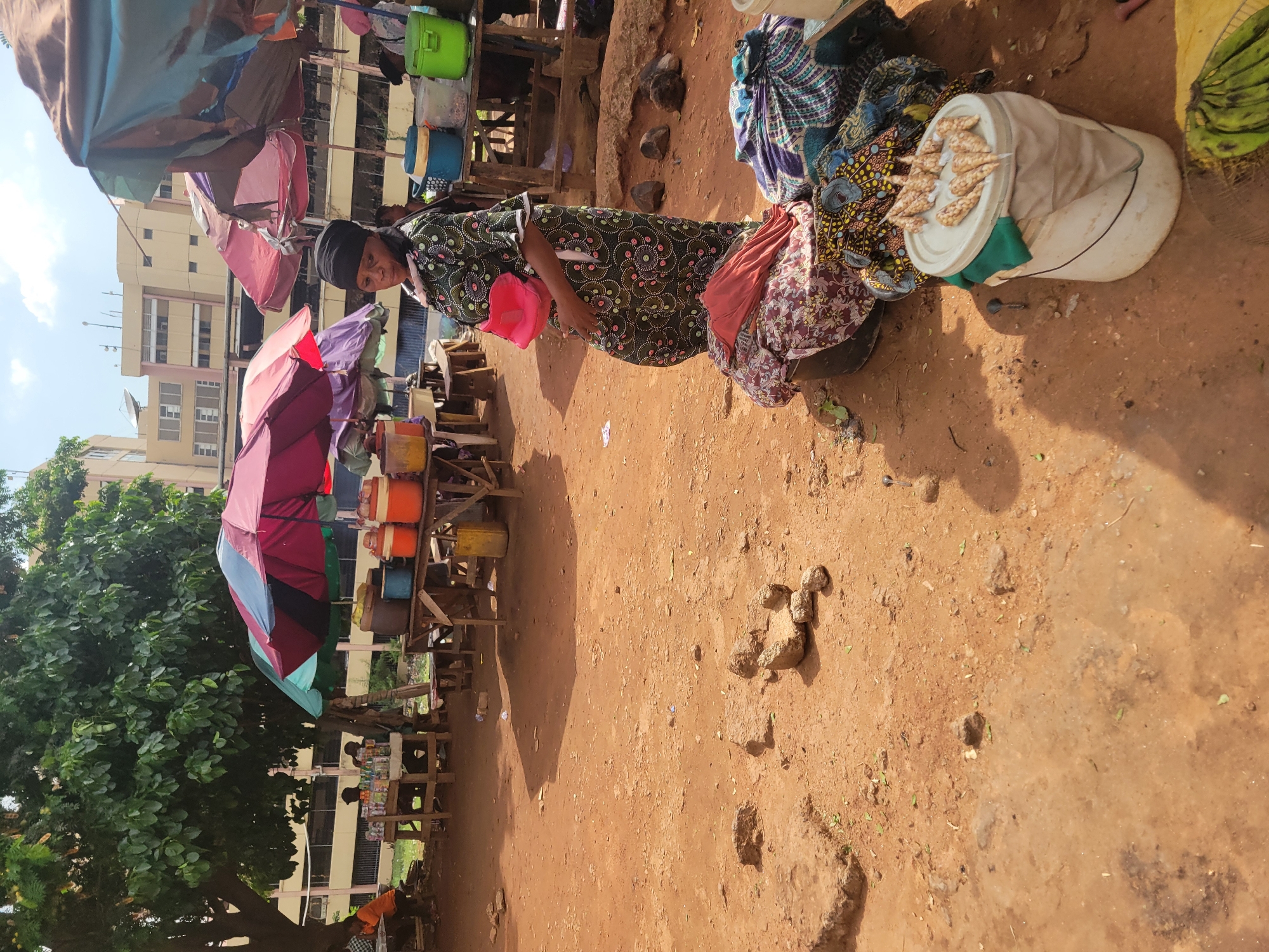 An outdoor scene in Abuja, Nigeria, shows a woman standing near a market stall on a dirt-covered ground. The woman, wearing a dark head covering and a patterned long dress, holds a pink bag. Beside her are sacks of clothes and a white bucket containing what appears to be food. Behind her, a row of stalls displays colorful containers, likely filled with goods for sale. An open umbrella provides shade to the stalls. In the background, a building is visible, and a tree with lush green foliage stands on the left side of the frame. The ground is reddish-brown with scattered rocks and debris.