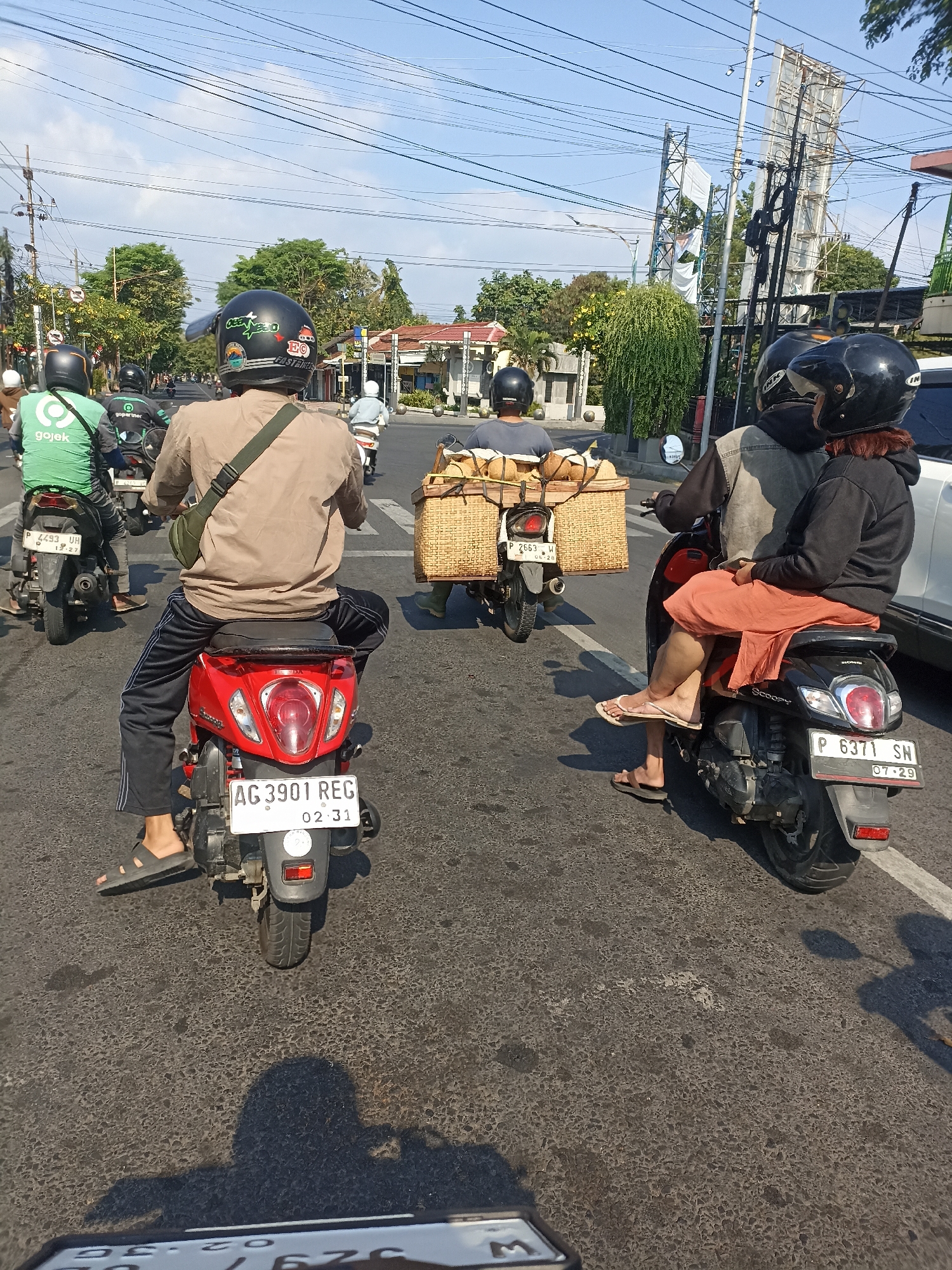 In Mojopanggung, Indonesia, on a sunny day, several motorcycles navigate a street. In the foreground, a person wearing a tan shirt and black pants rides a red scooter, their right foot resting on a black sandal. Behind them, a Gojek rider in a green jacket with the company logo is visible on their black motorcycle. To the right, two individuals on a black scooter are captured. The passenger, wearing an orange skirt and black hoodie, is seated behind the driver. The driver, wearing a gray vest over a dark shirt, is partially obscured. This scooter carries a large woven basket filled with coconuts. Further down the road, another motorcycle with a basket of coconuts is seen, along with other vehicles and pedestrians, suggesting moderate traffic. Power lines crisscross the bright blue sky, and lush green trees line the street, with buildings visible in the background. The road surface appears worn, with some patched areas and oil stains.