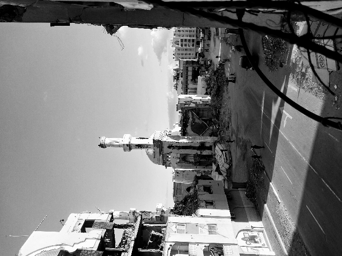 The black and white photograph shows a street heavily damaged by war. Buildings on either side are partially destroyed. A mosque with a prominent minaret stands in the middle, also bearing signs of damage. People are walking or standing in the street amidst the rubble. Debris is scattered everywhere. Overhead wires are visible. The sky is clear. The perspective is from a slightly elevated position, looking down the street.